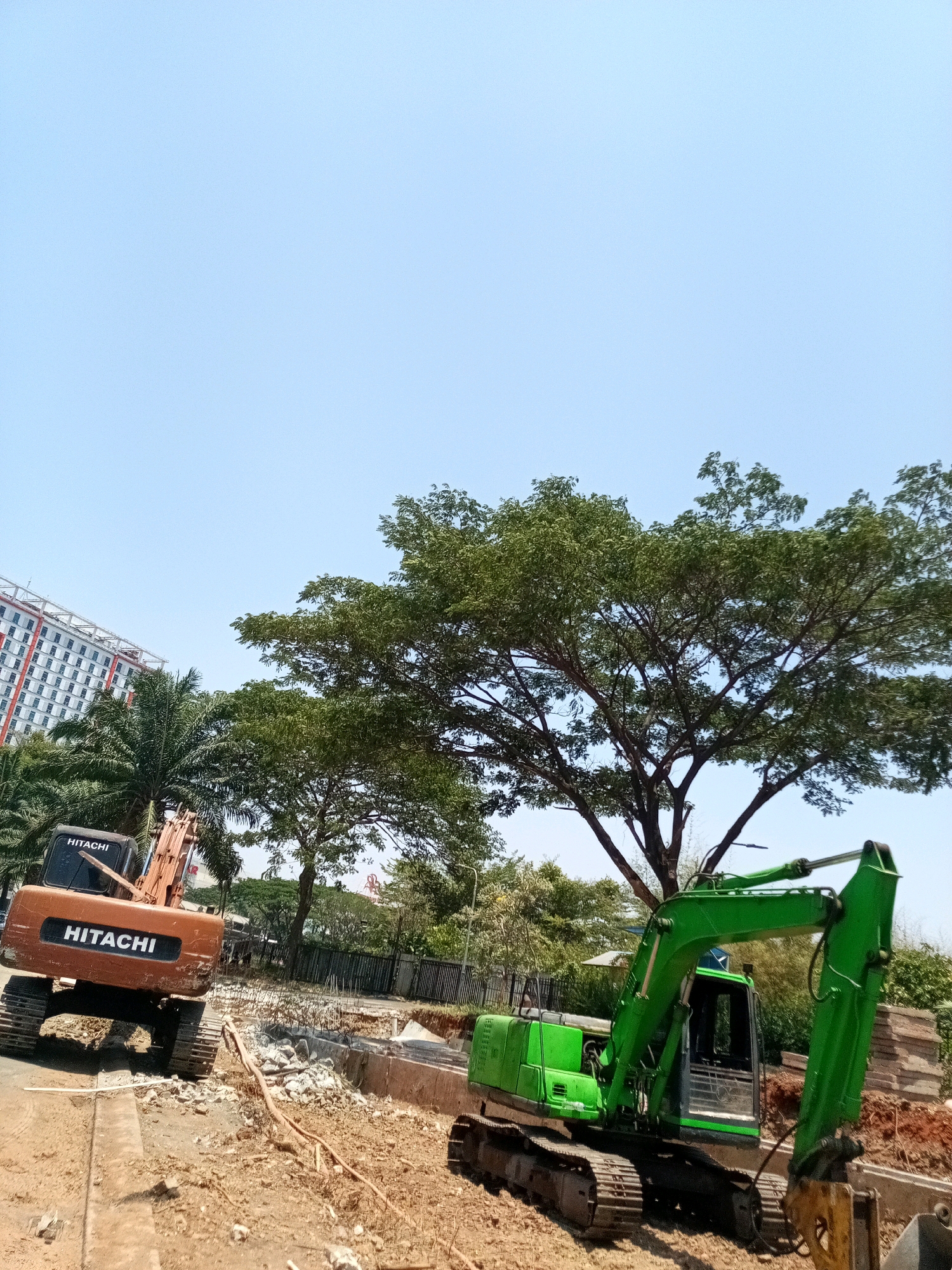 The image depicts a construction site under a clear blue sky in Bekasi, Indonesia. In the foreground, two excavators are present. The one on the left is orange and bears the "HITACHI" logo. To its right, a green excavator is positioned. The ground is covered in dirt and debris, indicative of ongoing construction or demolition work. In the background, a tall building with a red and white facade is visible, alongside lush green trees, suggesting an urban or semi-urban environment. The scene captures a moment of activity at a construction project, with heavy machinery ready for operation.