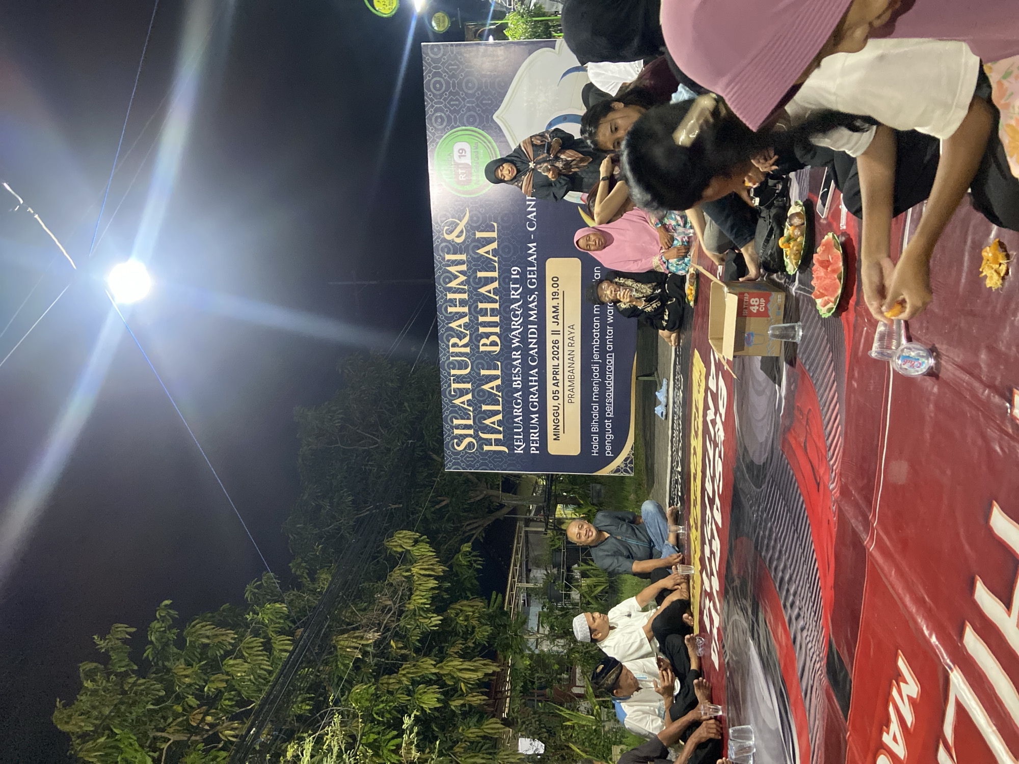 People are gathered at night for a "Silaturahmi & Halal Bihalal" event in Gelam, Indonesia. A banner indicates the event is for Keluarga Besar Warga RT 19 (a local neighborhood community) at Perum Graha Candi Mas, and took place on Sunday, April 5, 2026, at 7 PM, on Prambanan Raya street. People are sitting on a red mat, eating fruit and drinking water from plastic cups. Some sit cross-legged on the ground. Trees are in the background, and a bright light illuminates the area.