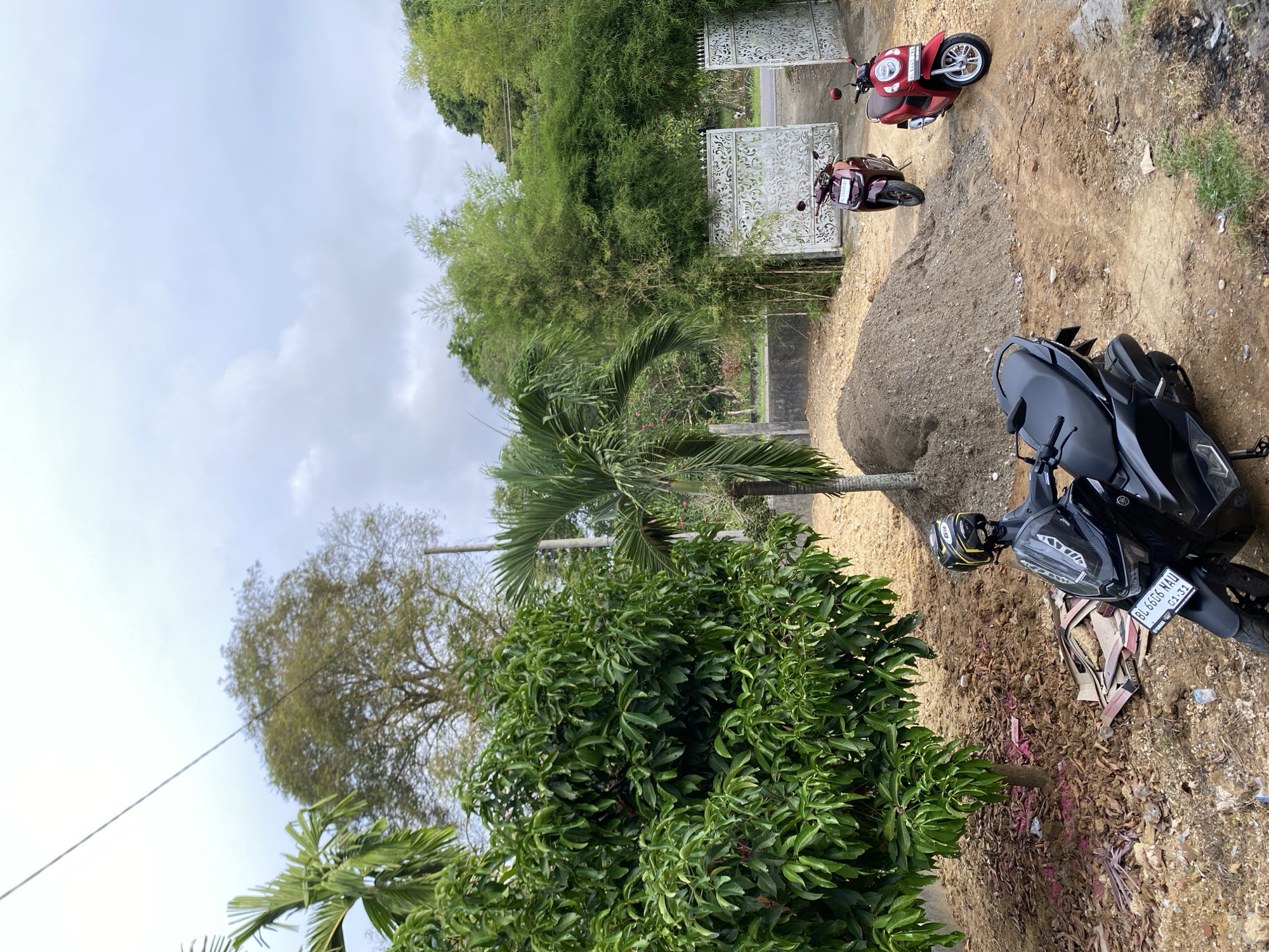 In the city of Meunasahlhok, Indonesia, this image captures an outdoor scene with a focus on parked motorcycles and lush vegetation. A black scooter, identified by the license plate "BL 6666 NAU", is positioned in the foreground on a dirt path, a yellow and black helmet resting on its seat. Behind it, a pile of construction soil is visible. Further back, two more scooters are parked near an ornate white gate, one maroon and the other red.
The environment is characterized by abundant greenery, including a large, leafy tree to the left, a palm tree, and bamboo stalks to the right. In the distance, hills covered in trees form the backdrop, under a cloudy sky. The ground is primarily unpaved, with patches of dry leaves and scattered debris.