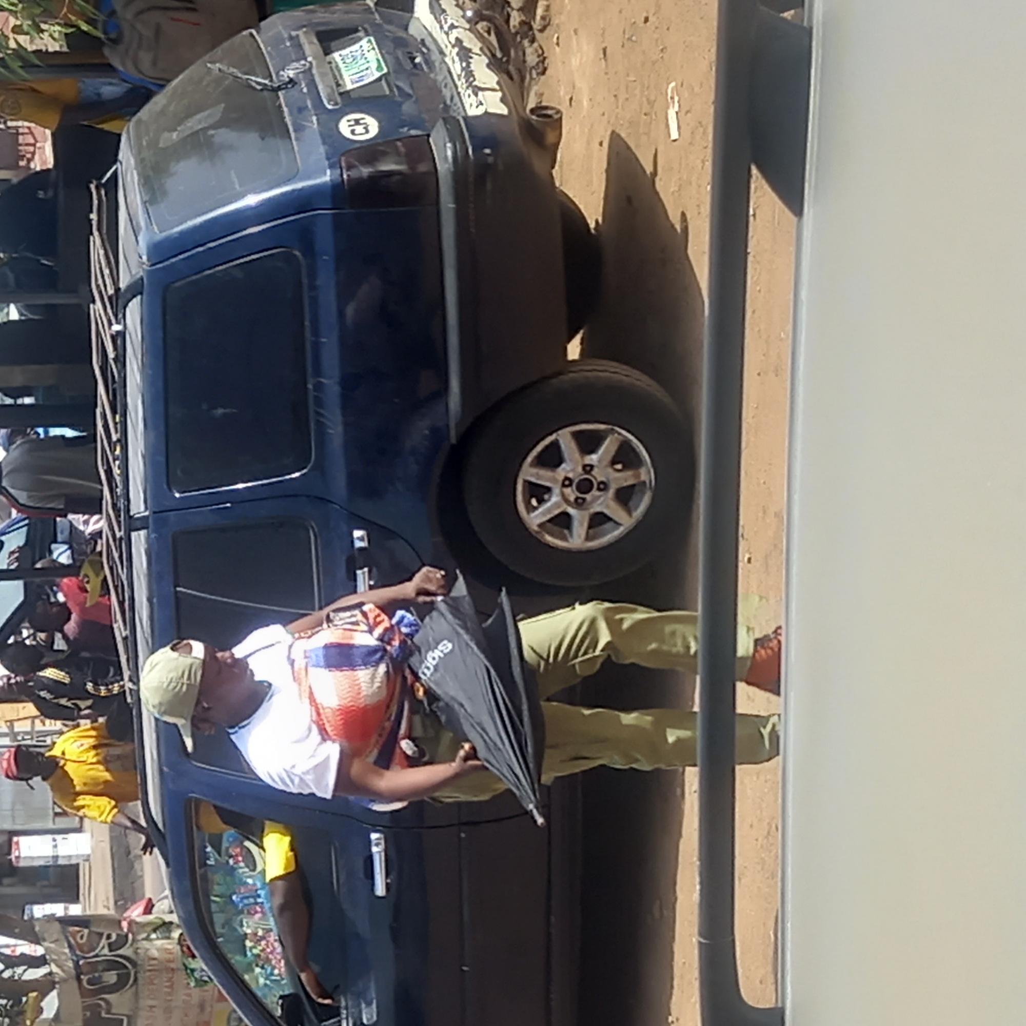 The image, taken in Dadi Riba, Nigeria, captures a bustling outdoor scene under bright daylight. The main subject is a dark blue, possibly older model, SUV or minivan-style vehicle. It features a roof rack laden with various bags and items, and a spare tire mounted on its rear. A white circular sticker with "HO" is affixed to the back of the vehicle, alongside a green sticker displaying some Arabic script, "Kano," and "R 20."
Standing beside the vehicle, partially leaning against its open front passenger door, is a man wearing a white t-shirt, light-colored (possibly beige) cap, and olive green trousers. He has a colorful woven bag/backpack slung over his shoulder and is holding a black, folded umbrella. The brand "SIGO" is visible on the umbrella. His stance suggests he might be waiting or interacting with someone.
Several other individuals are visible around and within the vehicle. One person in the vehicle's driver's seat appears to be wearing a yellow shirt, and another individual is visible in the background further left, also in a yellow shirt, with what looks like "POLICE" text partially visible. The setting appears to be a street or market area, characterized by a sandy or dusty ground. Shadows are cast by the vehicle and people, indicating a sunny day. The overall impression is that of a public transport or gathering point. On the far right, a tall, light-colored vertical structure partially obscures the scene.