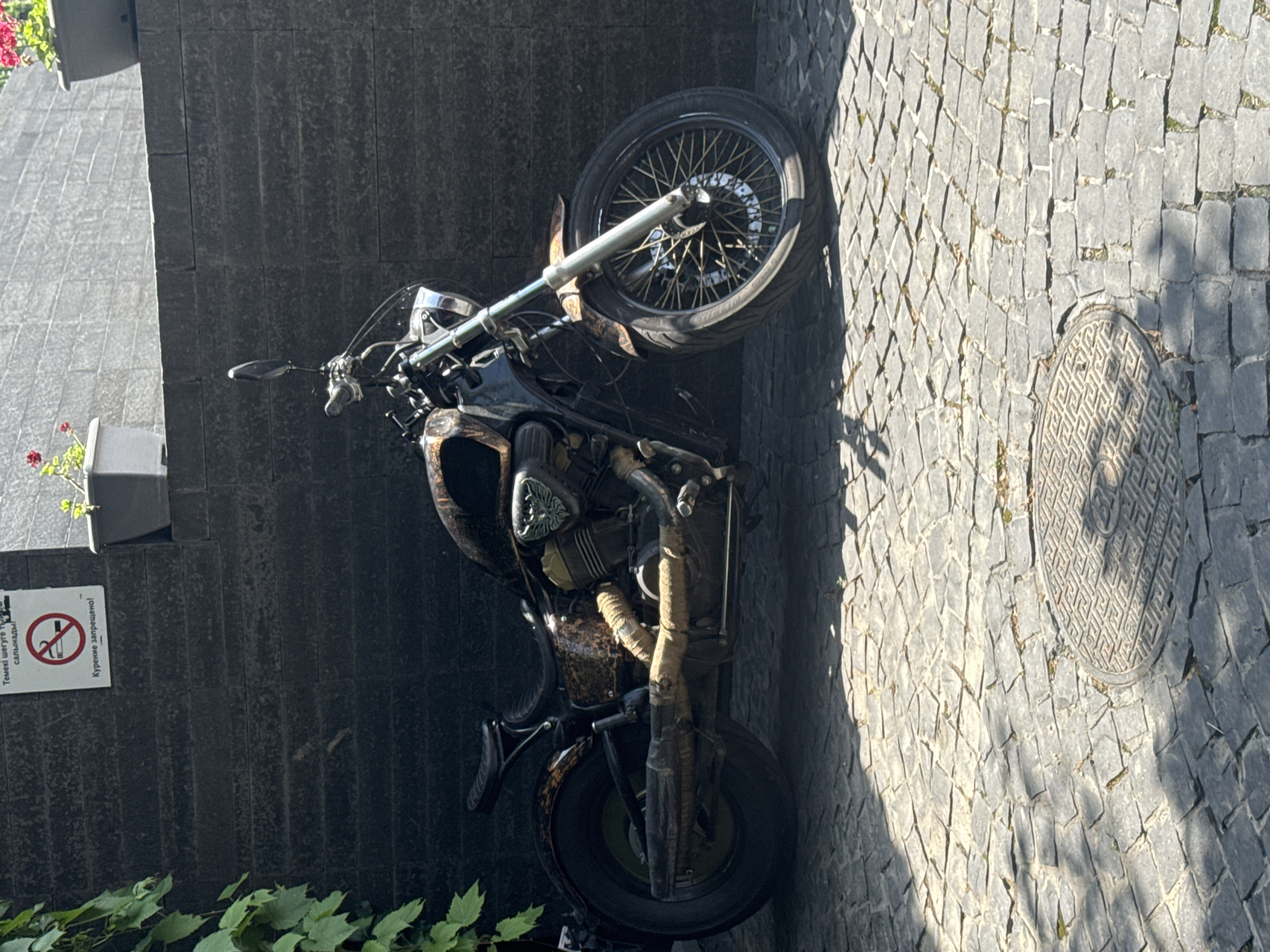 A vintage motorcycle is parked on a cobblestone street in Almaty, Kazakhstan. The motorcycle is black with chrome accents and appears to be in good condition, though it has a rustic, weathered look. It is positioned leaning against a dark, textured wall. In the background, there is a manhole cover with an intricate pattern. Sunlight casts shadows across the scene, suggesting it is daytime. A small sign is visible on the wall, with text in Cyrillic and what appears to be a no-smoking symbol. The overall atmosphere is one of urban street photography, highlighting a classic object in its environment. The vibrant green leaves of a plant are visible in the foreground, adding a touch of nature to the urban setting.