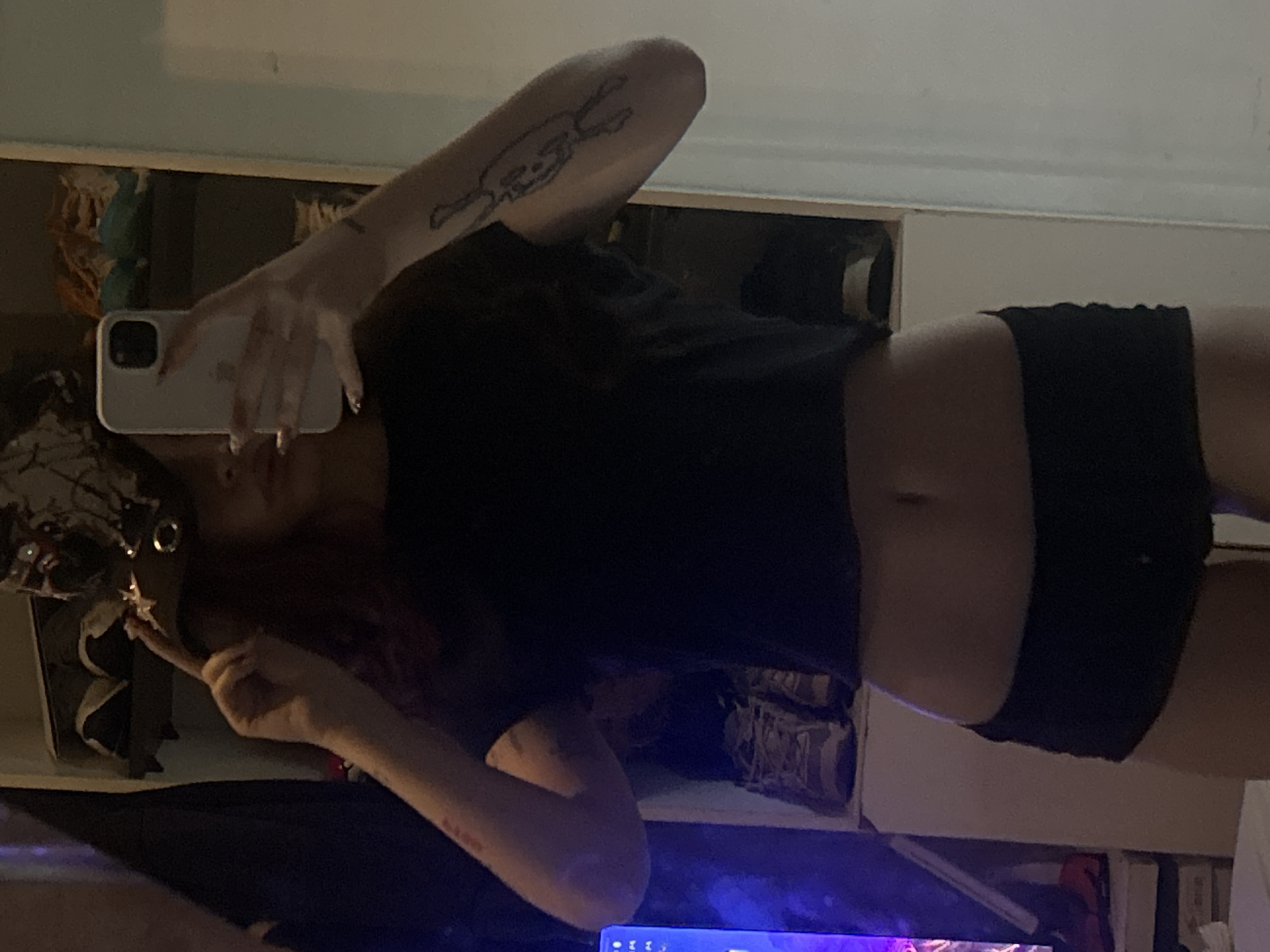 A person is taking a selfie in what appears to be a bedroom. The person is wearing a black cropped t-shirt and black shorts. The room is cluttered with various objects including shoes, a Christmas tree, and a computer. It is not possible to identify the location as Buenos Aires, Argentina.