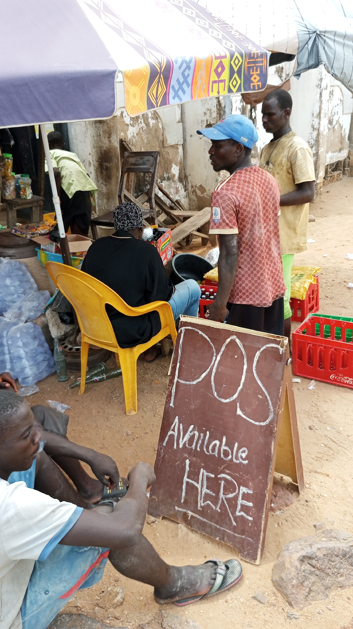 The image depicts an active outdoor street scene, likely a small marketplace or vendor stall, in Lokoja, Nigeria. A large, patterned sunshade provides cover over the area, which has a dirt ground.
In the foreground, a young man sits on the ground with his legs crossed, wearing a white t-shirt and blue shorts, focusing on something small in his hands. Next to him, a brown A-frame sign prominently displays "POS Available HERE" in white chalk.
Behind the sign, a man wearing a blue baseball cap and a red patterned t-shirt stands, looking towards the left. Behind him and slightly to the right, another man in a light yellow t-shirt stands. Further back, a person is seated on a bright yellow plastic chair, facing away from the viewer, wearing a black top and blue jeans. On the far left, another individual in a light green shirt is partially visible, bending over near a wooden stand with various jars.
Numerous objects suggest a retail or service point: several red Coca-Cola crates, other green plastic crates, plastic bags filled with water bottles or other items, and empty glass bottles on the ground. A small wooden table holds more items in jars. The general atmosphere suggests a busy, everyday commercial activity. The time of day appears to be bright daylight.