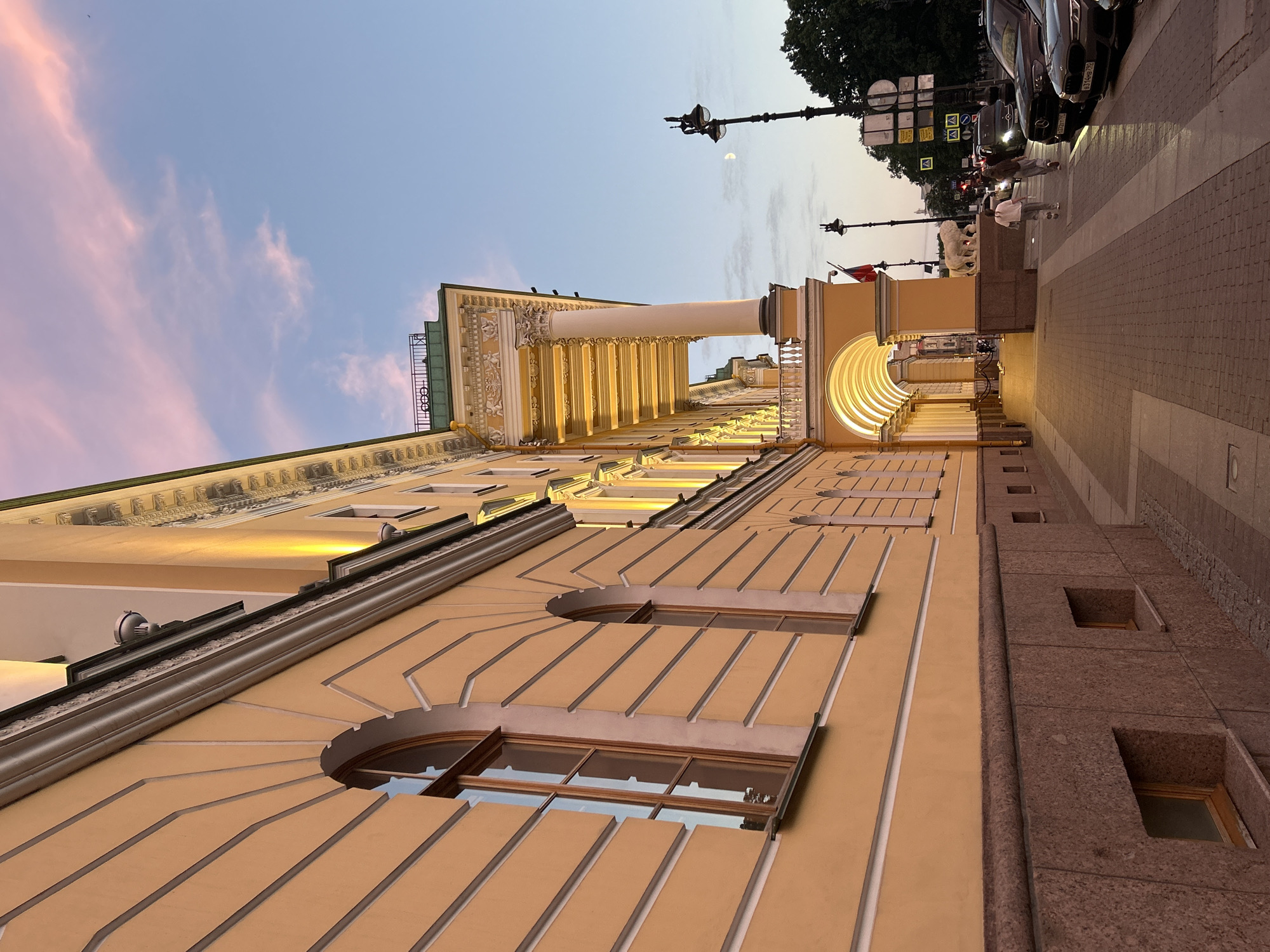 A low-angle, eye-level shot captures the side of a large, ornate yellow building with arched windows and a colonnaded walkway illuminated by warm lights. The building's facade features classical architectural elements, including decorative molding and columns. The sky above is a soft gradient of pink and blue, indicating either dawn or dusk, with wispy clouds. A street runs alongside the building, with a brick sidewalk. Cars are parked along the street. Two women walk away from the camera on the sidewalk. A lamppost with ornate detailing stands on the sidewalk, and the moon is visible in the sky. A white sculpture of a lion is situated near the building's entrance. Traffic lights and street signs are present along the street. The overall scene depicts an urban environment at twilight.