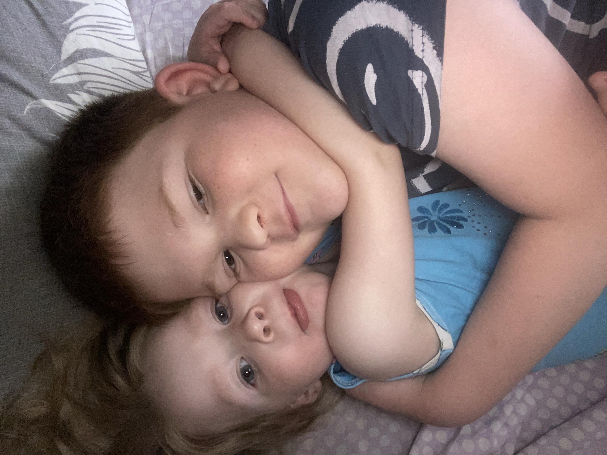 Two children, one with dark hair and one with blond hair, are lying on a patterned surface. The dark-haired child, wearing a striped shirt, has their arm around the blond-haired child who is wearing a light blue shirt. The location is unknown.