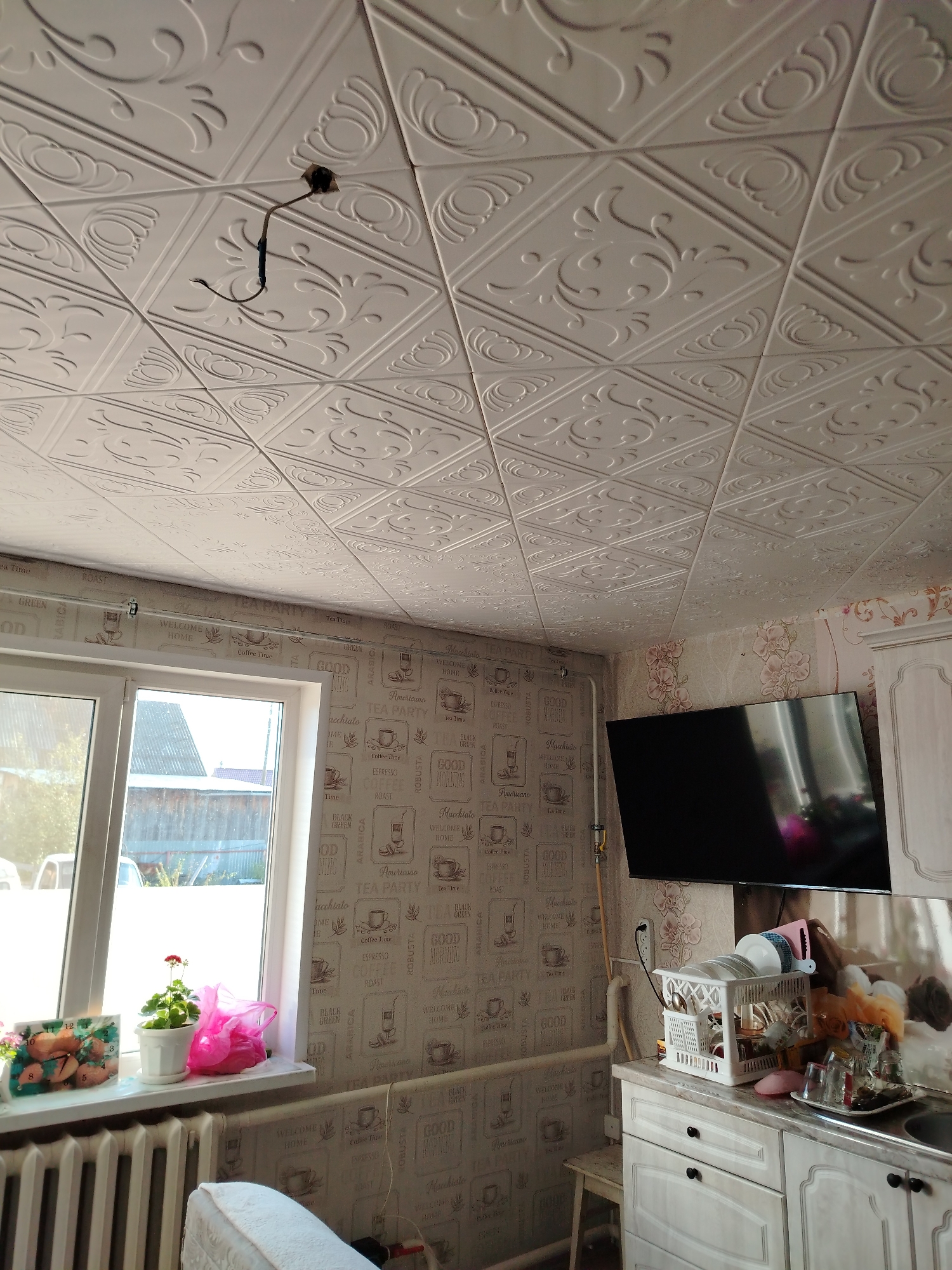 The image depicts an indoor scene, likely a kitchen or dining area, in a residential building located in Rusalevka, Russia. The ceiling is covered with decorative white tiles, each featuring a repeating embossed pattern of swirls and floral elements. A black electrical cable hangs loosely from the ceiling, connected to a conduit. The walls are covered with wallpaper that has a repeated pattern of coffee-related graphics and text, including phrases like "Tea Party," "Good Morning," "Coffee Time," and various coffee types like "Americano," "Espresso," and "Robusta."
To the left, a white-framed window reveals an outdoor view of a wooden fence and buildings, suggesting a residential neighborhood. On the windowsill below the window, there is a small potted plant with red flowers, a pink plastic bag, and a decorative clock displaying fruit. A white radiator runs along the base of the wall beneath the window.
On the right side of the image, a flat-screen television is mounted on the wall, reflecting the room's interior. Below the television is a countertop with kitchen cabinets. A white dish drying rack filled with dishes and cutlery sits on the countertop, along with several glasses on a tray. The cabinets have dark knobs and appear to be light-colored. A portion of a stool is visible in the lower right corner. The overall impression is of a lived-in domestic space.