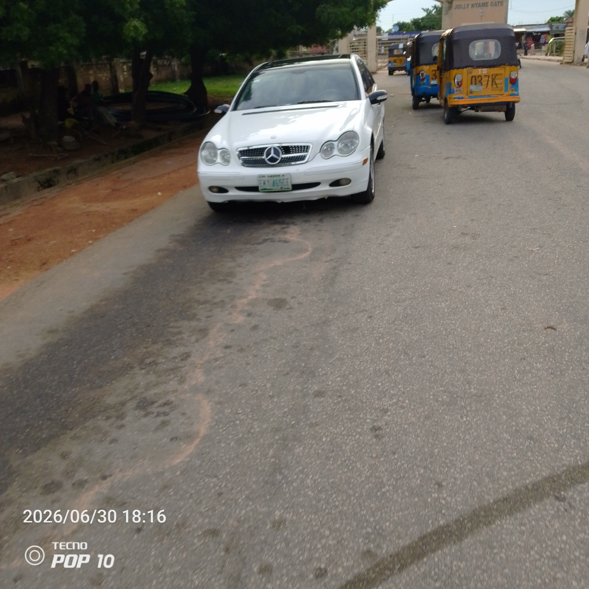 The image captures a street scene in Jalingo, Nigeria, featuring a white Mercedes-Benz sedan parked on the right side of the asphalt road. The car has a visible license plate that reads "FKY 465CE". In the background, two yellow and blue auto-rickshaws, also known as Keke Napeps, are visible, one behind the other. The front auto-rickshaw has "037K" on its license plate. Further in the background, a building with "JOLLY NYAME GATE" inscribed above it can be seen. Trees line the left side of the road, with some figures partially obscured by foliage. The road surface shows some wear and tear, with a distinctive water stain or reflection creating a wavy pattern in the foreground. The lower-left corner of the image displays a timestamp: "2026/06/30 18:16" and the TECNO POP 10 logo.