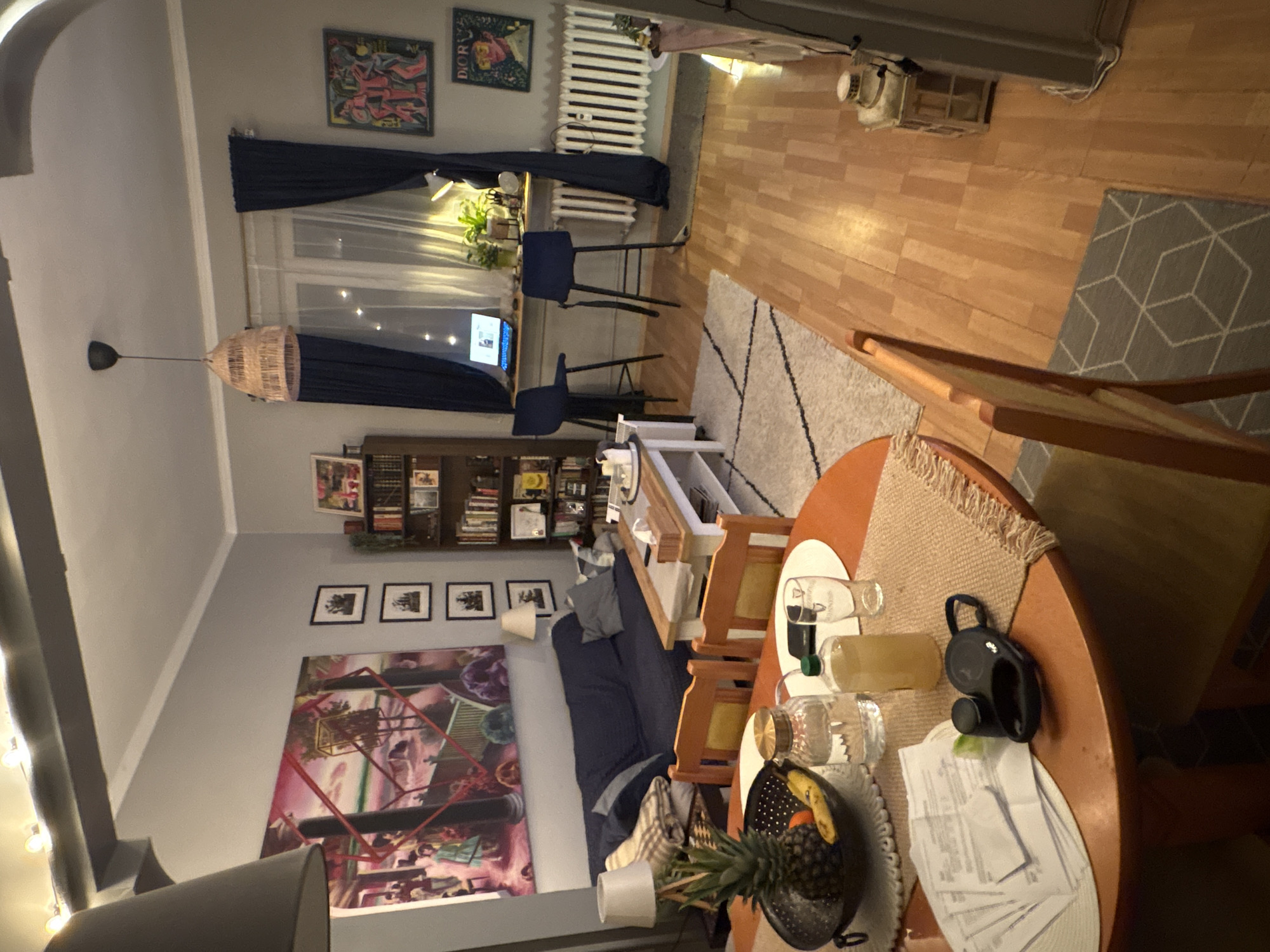 The image shows an interior space, likely an apartment, with a living area and a dining table in the foreground. The living area includes a blue sofa, a coffee table, and a bookshelf filled with books and decorative items. The dining table is set with glasses, bottles, a fruit bowl, and papers. There's a pineapple on the table. A geometric patterned rug is visible between the living area and what appears to be a workspace with two chairs and a desk near a window. The wall is decorated with several framed artworks and photographs. The lighting is warm, suggesting evening or artificial light. The city of Sofia, Bulgaria is not visually discernible from the image, so no inference can be made about the location.