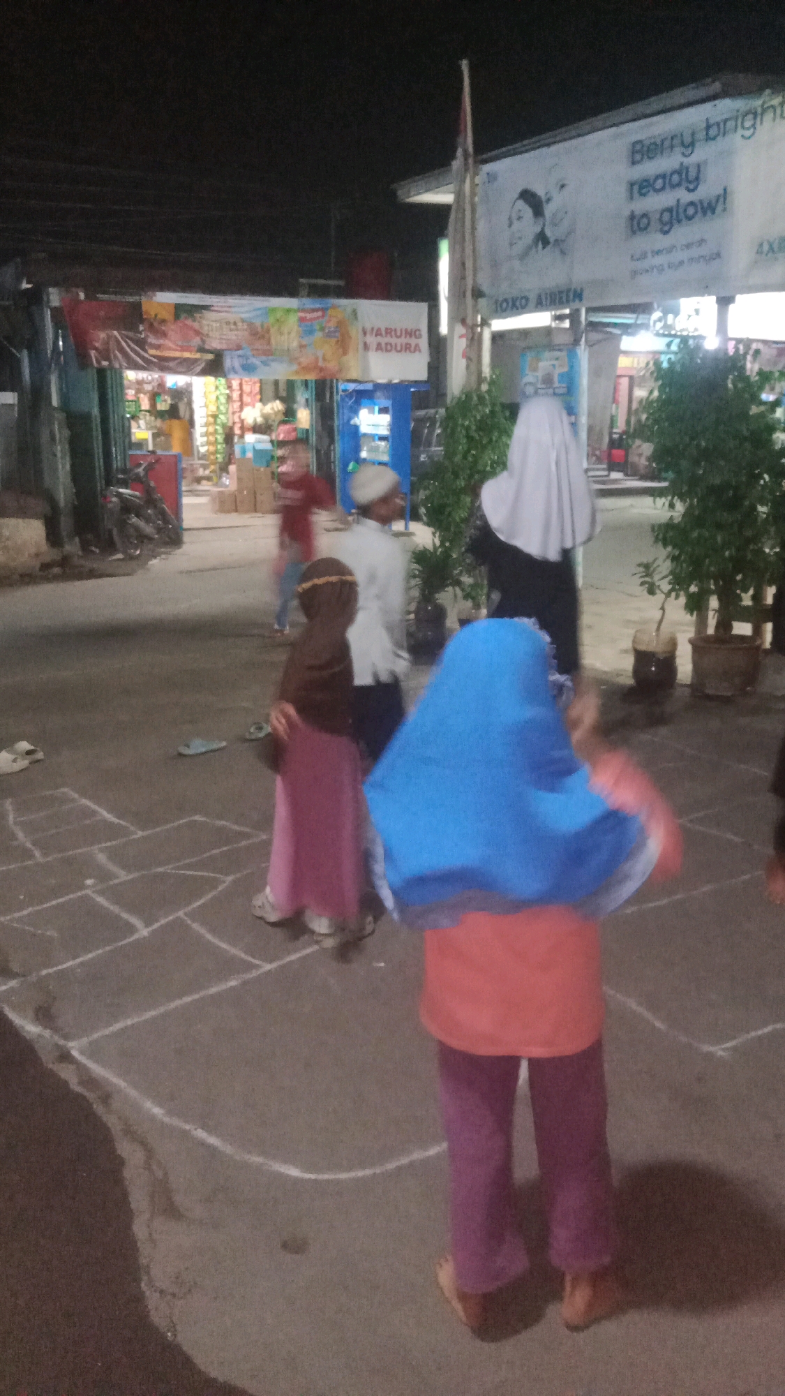 At night in Sumberjaya, Indonesia, several individuals are seen in an outdoor area that appears to be a street or open courtyard. The ground is paved, and hopscotch grids are drawn with white chalk. In the foreground, a child in a blue headscarf and an orange shirt is visible from the back, standing on a chalk square. To the left of this child, another child in a brown hijab and a pink dress stands facing away from the camera. Further back, two adults are present. One adult, wearing a white shirt and a white cap, is walking. Another adult is partially visible wearing a white hijab. In the background, there are storefronts with illuminated signs. One sign reads "WARUNG MADURA" and another appears to be an advertisement for "Berry bright ready to glow!". A motorcycle is parked to the left of the frame. The scene suggests a community gathering or children playing outdoors in the evening.