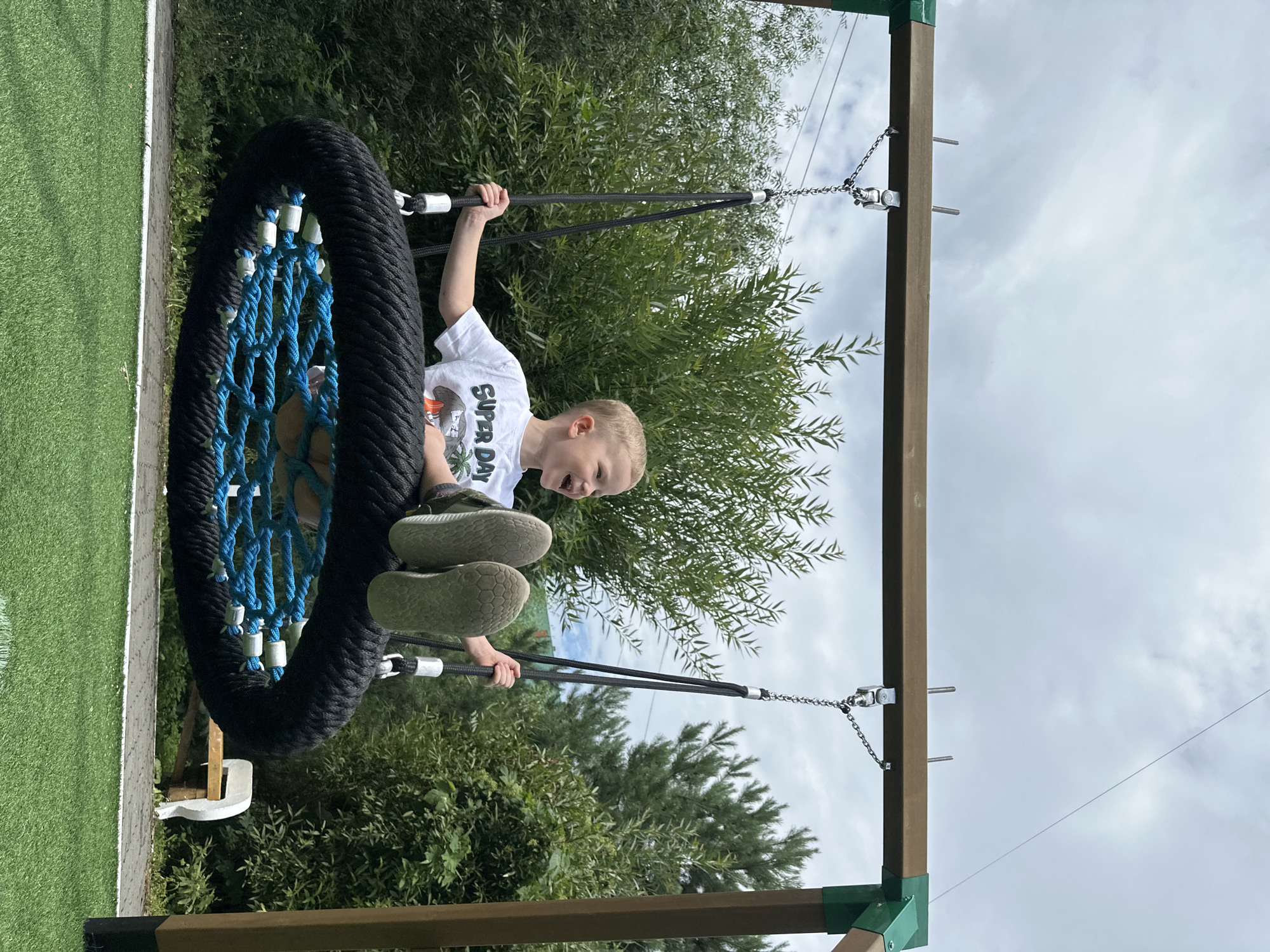 A young boy is joyfully swinging on a large, round, black-roped swing with a blue net in the center. He is wearing a white t-shirt with "SUPER DAY" printed on it and green sneakers. The swing set is made of wood with green metal accents. The background consists of lush green trees and a cloudy sky. The ground is covered with artificial green turf. This image appears to be taken in an outdoor park or playground, possibly in the city of Danilovka (Altai), Russia.