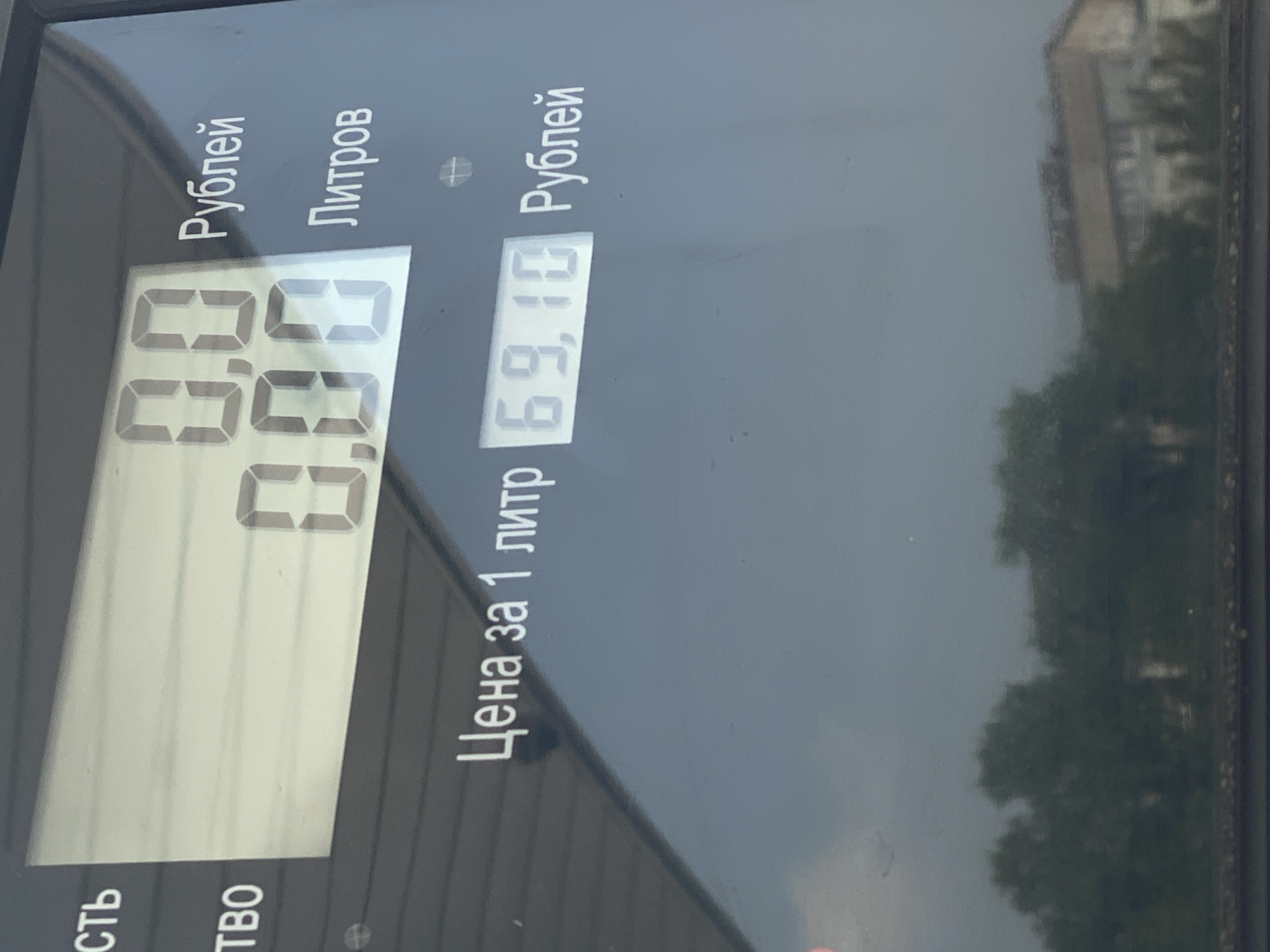 The image is a close-up shot of a digital display on what appears to be a gas pump. The display shows the numbers "0.0" for the quantity dispensed and "0.00" for the total cost, both in rubles. Below these figures, it indicates "Цена за 1 литр" (price per 1 liter) followed by "69,10" rubles. The background of the image is a reflective surface showing a hazy, overcast sky and the blurred outlines of trees and a building, suggesting an outdoor urban or suburban environment. Based on the Cyrillic text and the building style, the location is identified as Novokuznetsk, Russia.