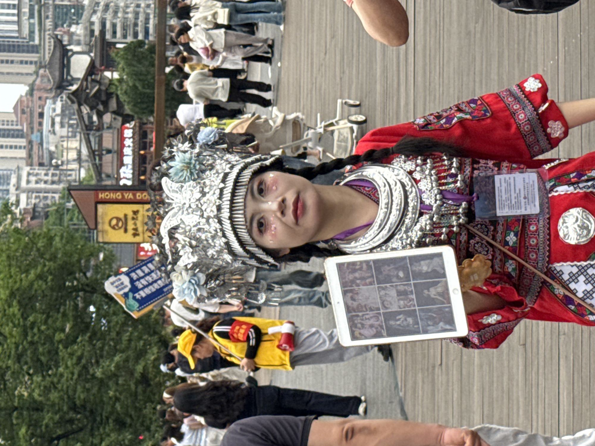 A woman wearing a traditional Chinese garment and headwear stands on a wooden deck, holding a tablet displaying images. She has a braid and makeup on her face. In the background are buildings and people, suggesting an urban environment. A sign reading "HONG YA DO" is visible. Other people are standing or walking nearby. It is daytime. The location is identifiable as Yuzhong District, China, based on the sign "HONG YA DO".