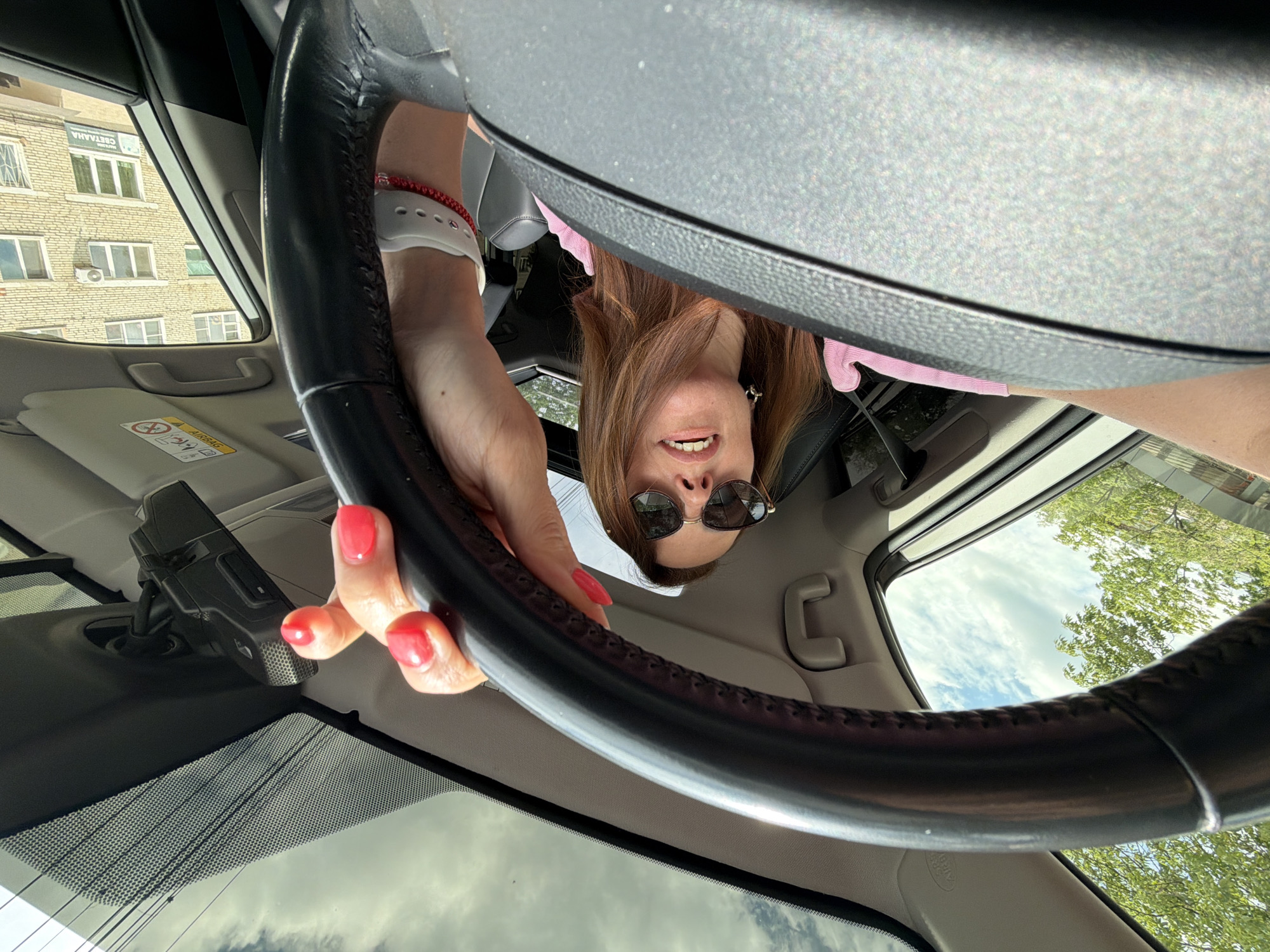 A woman is driving a car on a street in Birobidzhan, Russia. The woman has long brown hair, is wearing sunglasses, and a pink shirt. She is holding the steering wheel with her right hand. Her nails are painted red. In the background, there is a building and trees. The sky is visible through the sunroof of the car.