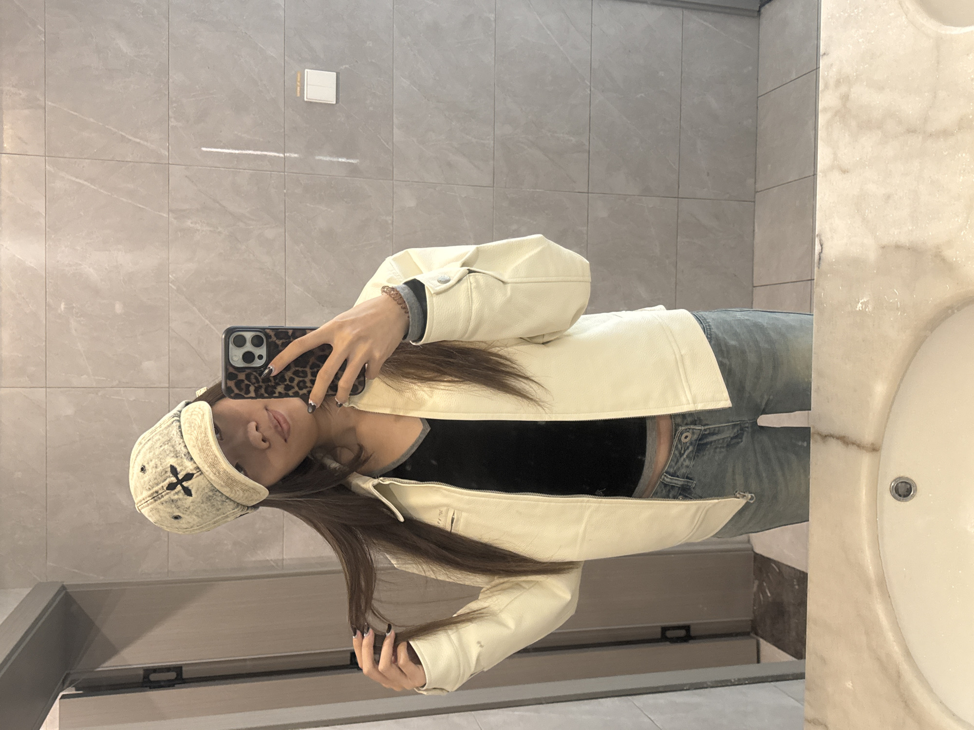A young woman is taking a selfie in a bathroom with light gray marble tiles and a white marble sink. She is wearing a beige denim hat with a black cross, a black top, a cream-colored leather jacket, and light blue jeans. She is holding a smartphone with a leopard-print case. The bathroom appears to be in a modern building in Nan'an District, China. A light switch is visible on the wall behind her.