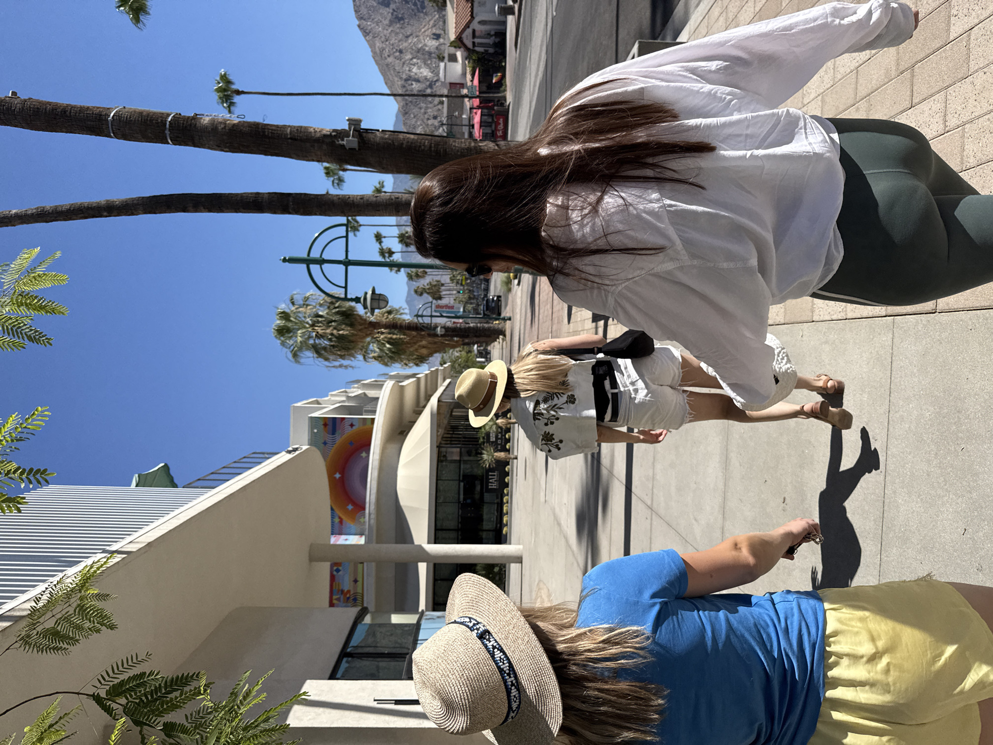 Three individuals are walking on a sidewalk on a sunny day in Palm Springs, California. The person on the left is wearing a blue t-shirt and yellow shorts, with their back to the camera. The middle person is wearing a white embroidered jacket and white shorts. The person on the right is wearing a white long-sleeved shirt and green leggings. In the background, there are palm trees, buildings with modern architecture, a street with cars, and mountains in the distance. One of the buildings features a colorful mural.