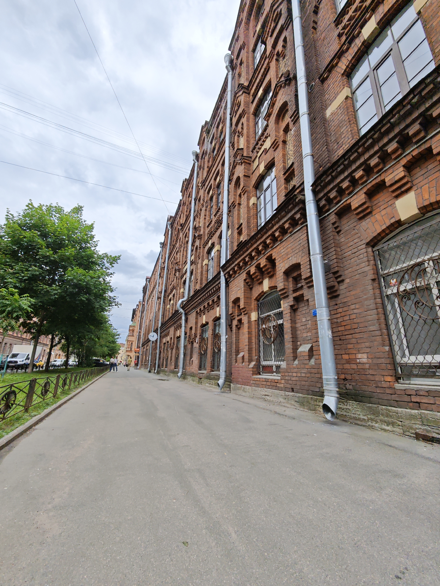 The image shows a street scene in Saint Petersburg, Russia, on a cloudy day. The perspective is low-angle, looking up at a tall, red brick building that occupies the right side of the frame. The building features ornate brickwork detailing, numerous windows with decorative metal grates, and prominent silver downspouts running vertically along its facade.
To the left of the building, a paved sidewalk runs parallel to it, with a line of mature green trees bordering the sidewalk and a decorative metal fence separating the sidewalk from the street. Several vehicles, including a white van and a car, are parked along the street. A few pedestrians are visible walking on the sidewalk in the distance, appearing as small figures.
The sky is overcast with gray clouds, suggesting a typical Saint Petersburg weather. The overall impression is an urban street with historical architecture and moderate pedestrian activity.