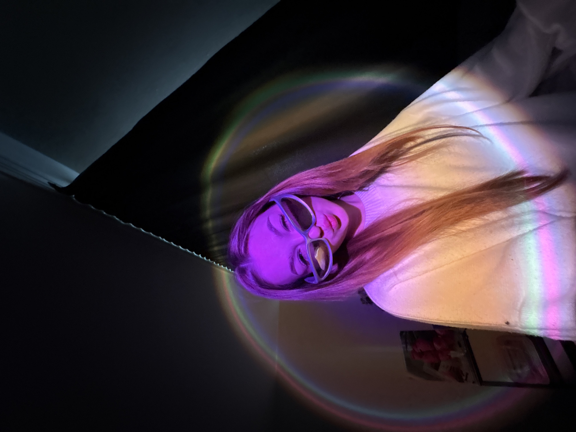 An indoor, close-up shot captures a person, likely female, lying horizontally within the frame, illuminated by projected rainbow light patterns. The person has light brown or auburn hair and is wearing a white top or blanket. Their face is largely cast in a strong purple hue from the light, with their eyes closed or heavily lidded, and lips slightly parted with pink lipstick. They wear large, clear-framed glasses that reflect some of the purple light.
A vibrant, circular rainbow light projection arcs across the upper body of the person, the white garment they are wearing, and the dark background. The rainbow colors (red, orange, yellow, green, blue, purple) are distinctly visible. The dark background, possibly a wall or ceiling, takes up the upper left and central portions of the image, contrasting sharply with the illuminated areas. Below the person's right arm, a small, framed object or picture is partially visible. The overall lighting suggests a dark room, likely evening or night, with the primary illumination coming from the artificial light source creating the rainbow effect. No text is visible in the image. The scene conveys a relaxed, contemplative, or artistic mood.
Location context: Chaikovsky, Russia.