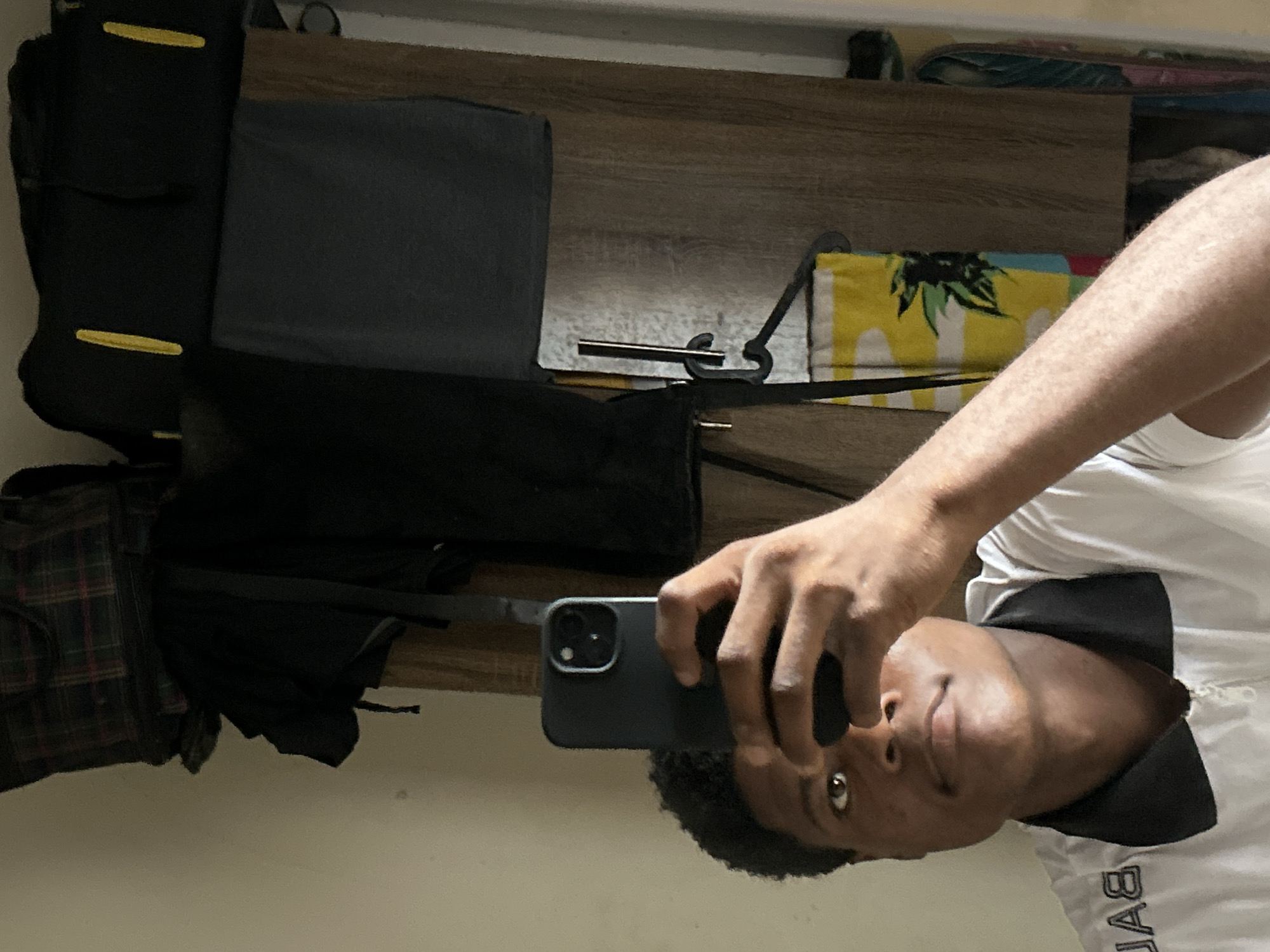 A dark-complexioned male, consistent with a young adult, is depicted from the chest up in what appears to be a mirror selfie. He holds a dark-cased smartphone in his raised right hand, obscuring part of his face. He has short, dark, curly hair and brown eyes. He is wearing a white polo shirt with a black collar and a visible zipper closure, with the letters "BA" visible on his left shoulder area. The background, seen as a reflection, consists of a plain light-colored wall on the left and a wooden-textured surface, possibly a wardrobe or door, on the right. Various items are visible in the background: a dark plaid bag and another dark bag with two yellow vertical accents are positioned at the top. Below these, dark clothing is present. A colorful towel, primarily yellow with green leaf patterns, hangs from a black hanger against the wooden surface. The scene represents an indoor environment located in Ihiagwa, Nigeria.