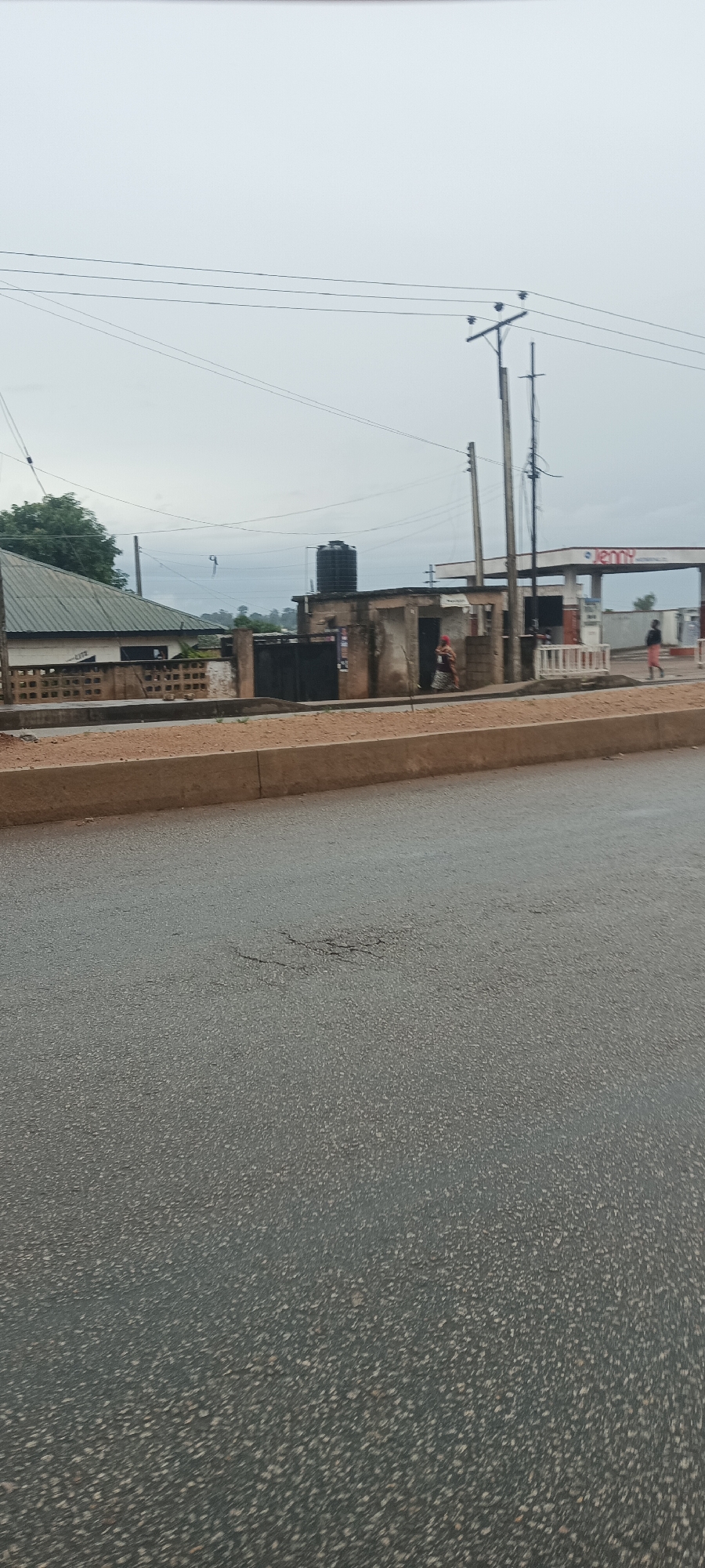The image depicts an overcast street scene in Bukuru, Nigeria, characterized by an urban-to-semi-urban environment. In the foreground, an asphalt road with visible cracks occupies the lower half of the frame. A low, reddish-brown concrete curb or median runs along the right edge of the road, separating it from the unpaved area beyond.
In the midground, several structures and elements are present. To the left, a building with a green corrugated metal roof and white walls is partially seen, flanked by green foliage. Adjacent to it stands a smaller, weathered concrete structure topped with a black water tank. Further to the right, a gas station canopy is visible, featuring partially legible signage that reads "JENNY PETROLEUM CO." Multiple utility poles, made of both wood and concrete, line the street and support a network of overhead power lines that crisscross the sky.
Two people are discernible in the distance on the right side of the road, walking towards the right. One individual is closer to the center, appearing to wear a red top, while another person in lighter clothing is seen near the gas station. The sky is uniformly overcast, providing soft, diffused lighting, and the road surface appears slightly damp, hinting at recent rain or high humidity. The scene portrays a routine daytime street view, marked by everyday infrastructure and pedestrian activity.