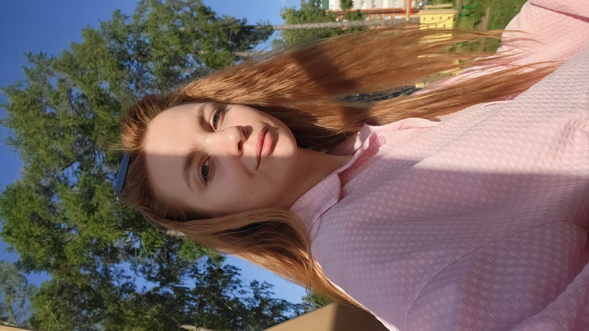 A woman with red hair and a light pink shirt poses for a selfie outdoors. She is standing in Chaikovsky, Russia. Behind her is a green tree and a blue sky. In the background is a building. The lighting is bright and sunny. Part of her face is shadowed.