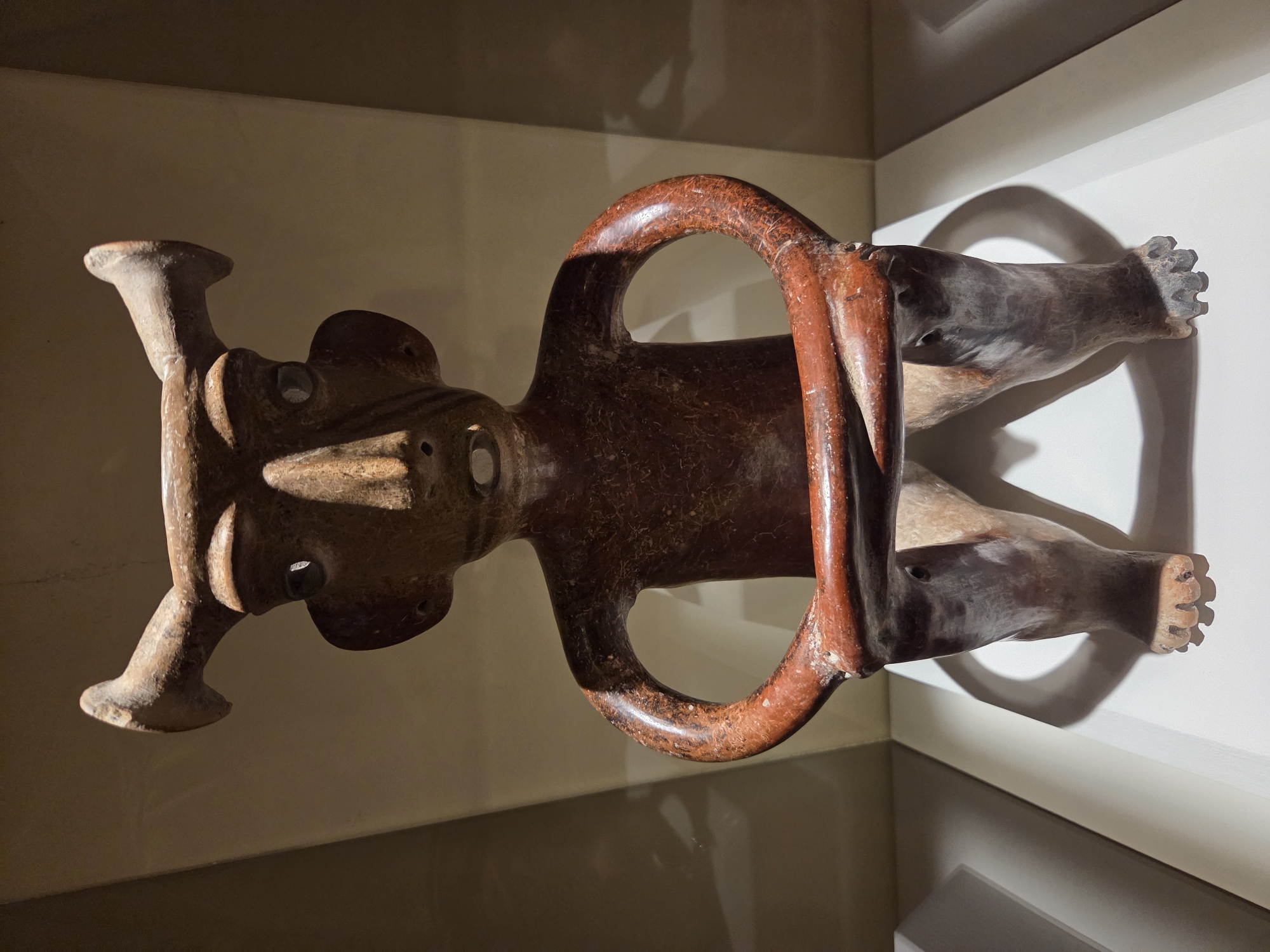 The image shows a full-front view of a seated anthropomorphic ceramic figure, likely an artifact on display. The figure has a prominent, long nose, wide-set eyes, and a mouth that appears open. It possesses two distinctive, curved horns extending from the top of its head. The figure's arms are bent, and its hands rest on its lap, clutching what appears to be a crossbar or a similar object. The body of the figure is reddish-brown with a textured surface, while the legs are a lighter, more muted color. The figure is positioned on a white surface, casting a shadow behind it against a neutral, dimly lit background. The context of Santa Ana, United States of America, is not directly discernible from the visual information presented in the image.
