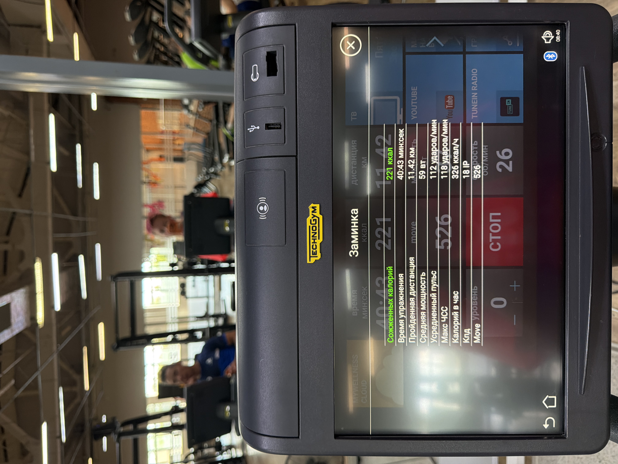 The image displays a close-up of a Technogym exercise machine console in a dimly lit gym. The console screen shows various workout metrics in Russian, including time, distance, calories burned, heart rate, and power output. The time displayed is 08:40. In the background, out of focus, are several people using other exercise equipment. The overall impression is of a typical gym environment. The location is identified as Chaikovsky, Russia.