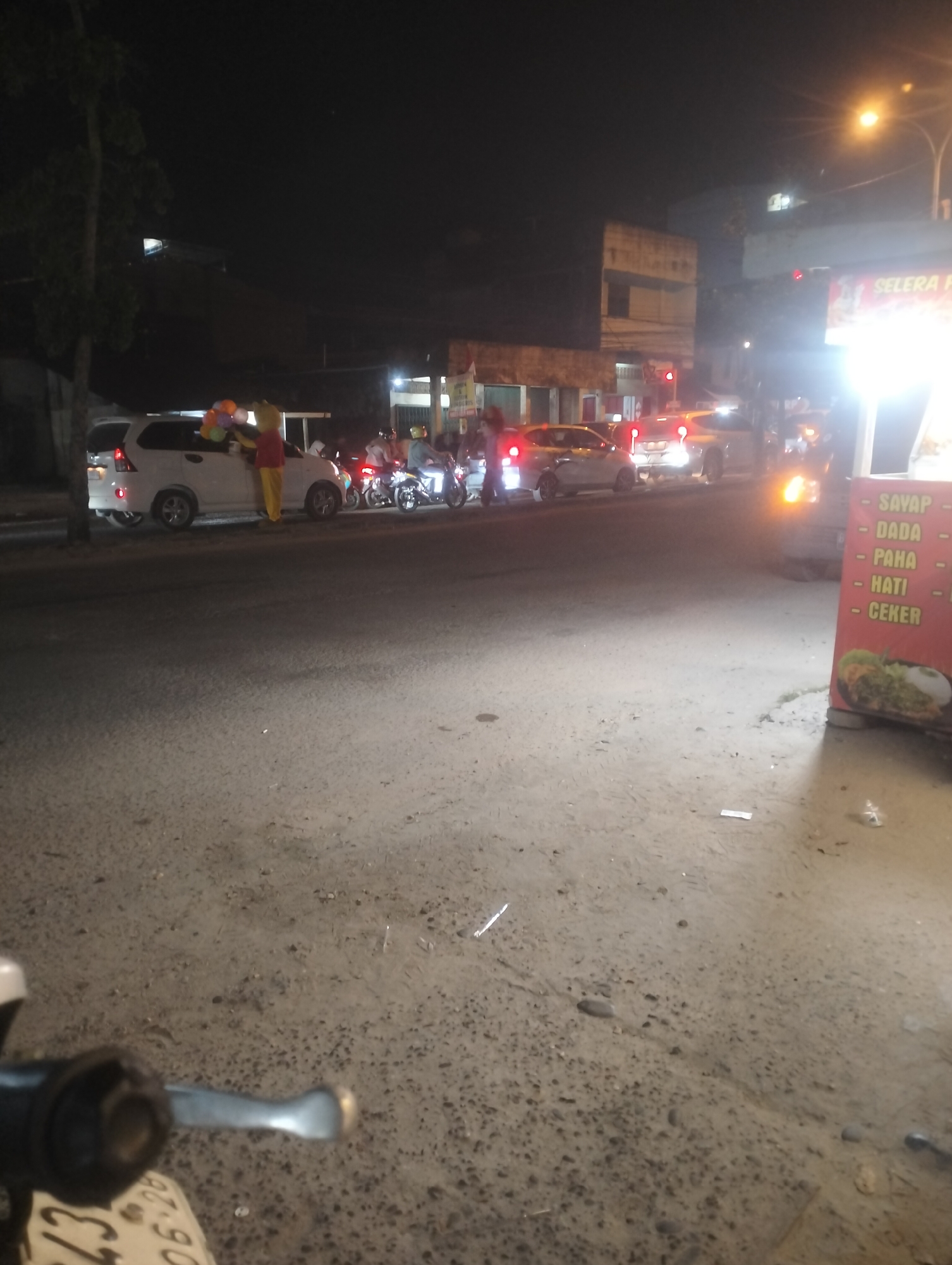 At night, in the city of Air Jamban, Indonesia, a street scene unfolds with traffic and a person dressed as a cartoon character holding balloons. The scene is illuminated by streetlights and the headlights of vehicles. In the foreground, a section of a motorcycle's handlebars is visible. A white vehicle, possibly an SUV, is parked on the side of the road, with a person in a yellow outfit and a red shirt, resembling a cartoon character, standing next to it and holding a bunch of colorful balloons. Several motorcycles are also present, some with riders. A red traffic light is visible in the background. A bright, illuminated sign on the right side of the image displays text that appears to be a menu, with items such as "SAYAP", "DADA", "PAHA", "HATI", and "CEKER". The ground is paved and appears to be a road surface. The overall atmosphere suggests a busy street at night with some form of promotion or event taking place.