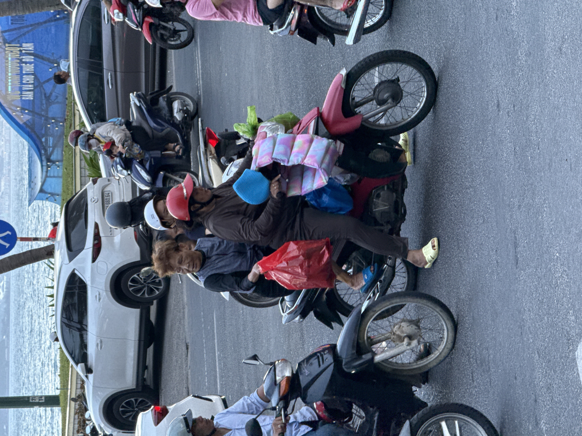 The image depicts a street scene in Nha Trang Ward, Vietnam, with multiple motorcycles and cars on a paved road. In the foreground, a person in a black shirt and brown pants is riding a pink motorcycle. They are wearing a red helmet and a blue head covering, and they are holding a padded blanket with pastel colors. To the left, another motorcycle, primarily black, is partially visible with a person wearing a light blue shirt and a helmet. Further to the left, a white SUV is parked.
In the middle ground, a woman with reddish-brown, permed hair is seated on a motorcycle, wearing a dark vest over a lighter top and holding a red plastic bag. She is wearing blue sandals. Beside her, another person, partially obscured, is seated behind her.
In the background, several other motorcycles and cars are present. One motorcycle is blue and carries two passengers, one of whom appears to be a child. A grey sedan is also visible. Across the street, a body of water, likely the sea, is visible, with a blue structure or awning in the distance. A traffic sign with the number "31 T4" is mounted on a pole on the left side of the image. The overall scene appears to be a busy urban environment during the day.