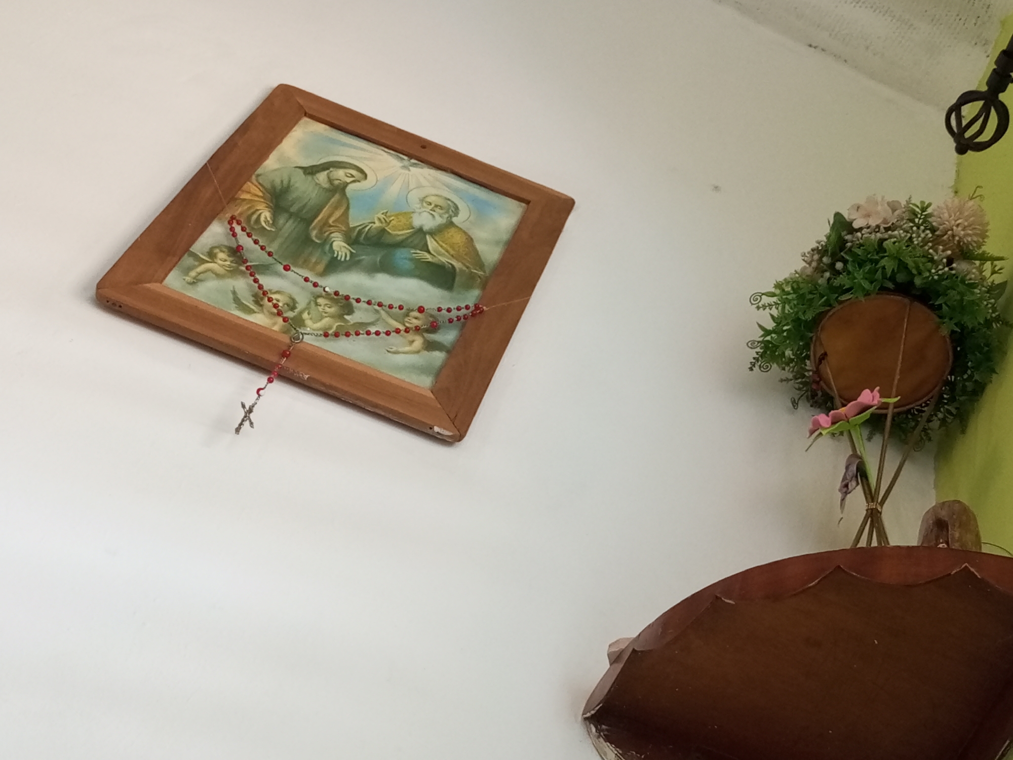 A framed religious painting hangs on a white wall. The painting depicts three figures and appears to be a religious scene. A rosary with red beads hangs from the painting, with a small cross visible. To the right of the painting, a floral arrangement is displayed, featuring green foliage and pink and white flowers. Below the floral arrangement, a portion of a dark wooden object, possibly furniture, is visible. The overall impression is a domestic or devotional setting. The location in the city of La Tagua, Venezuela, is not discernible from the image itself.