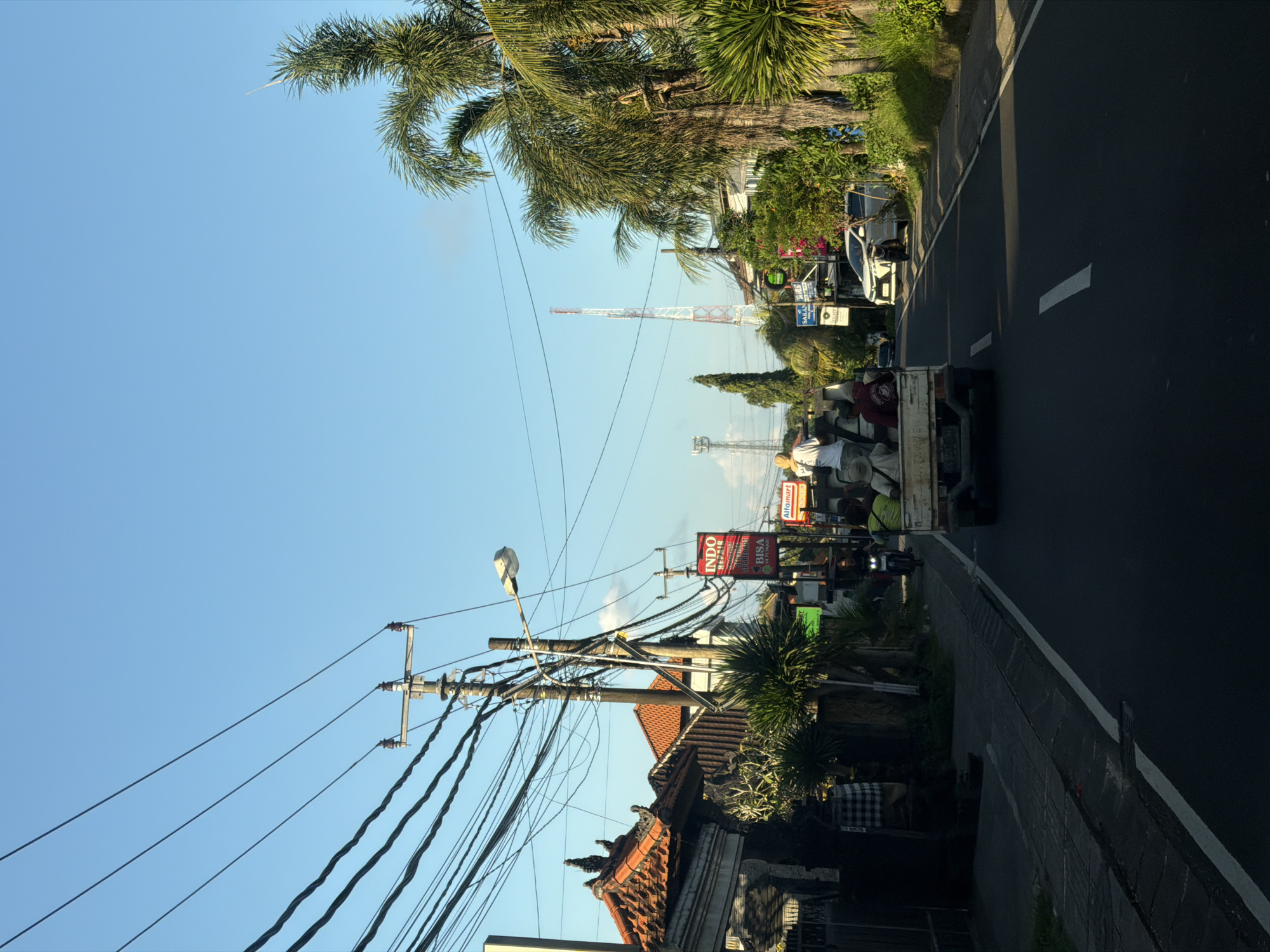 The image depicts a street scene in Kutuh, Indonesia, taken during daylight hours under a clear blue sky. The photograph is oriented vertically. A two-lane asphalt road with a white painted center divider runs from the bottom right toward the center of the frame. A white pickup truck with several passengers in the back is traveling on the road. Overhead power lines are heavily bundled and crisscross above the road. Palm trees and other vegetation are visible along the roadside. Buildings with traditional Balinese architecture, including tiled roofs, are located adjacent to the road. In the background, a tall, slender telecommunications tower is visible, with another slightly more distant, in the center of the frame. Various signs and advertisements, including one for "Indo BISA," are posted along the roadside. A streetlight on a wooden pole is visible on the left side of the road.