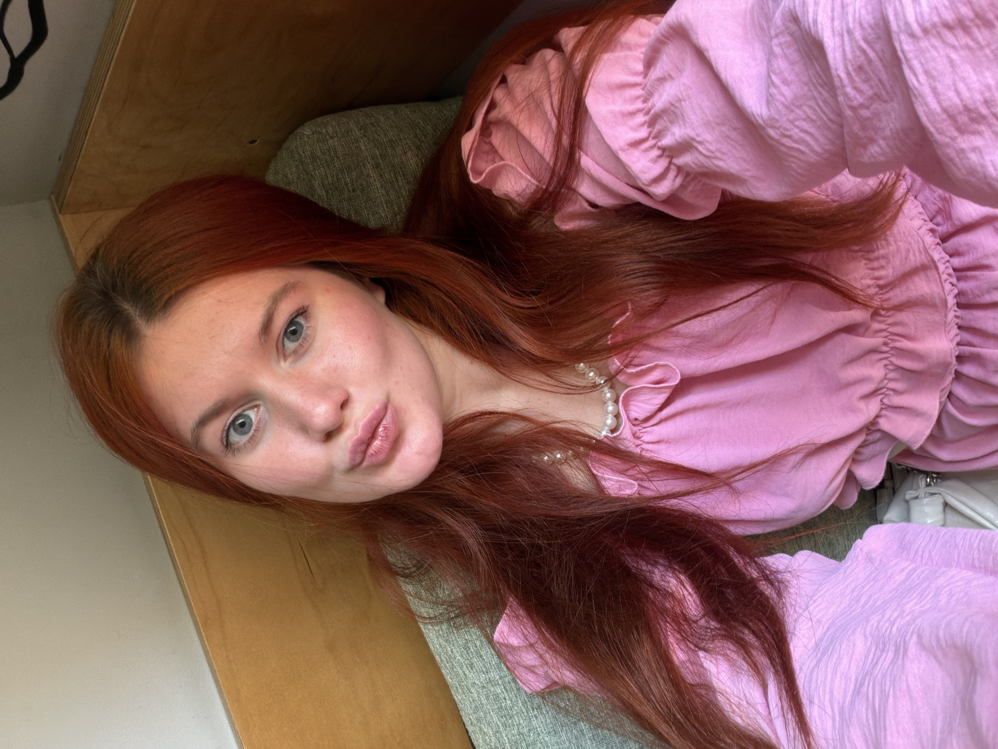 A young woman with long, red hair and blue eyes is captured in a close-up, selfie-style shot. She is wearing a pink dress with ruffled sleeves and a pearl necklace. Her lips are pursed in a kissing gesture. The background appears to be an indoor setting, possibly a room with wooden paneling and a textured green chair. There is no visible indication of the location being in Estosadok, Russia.