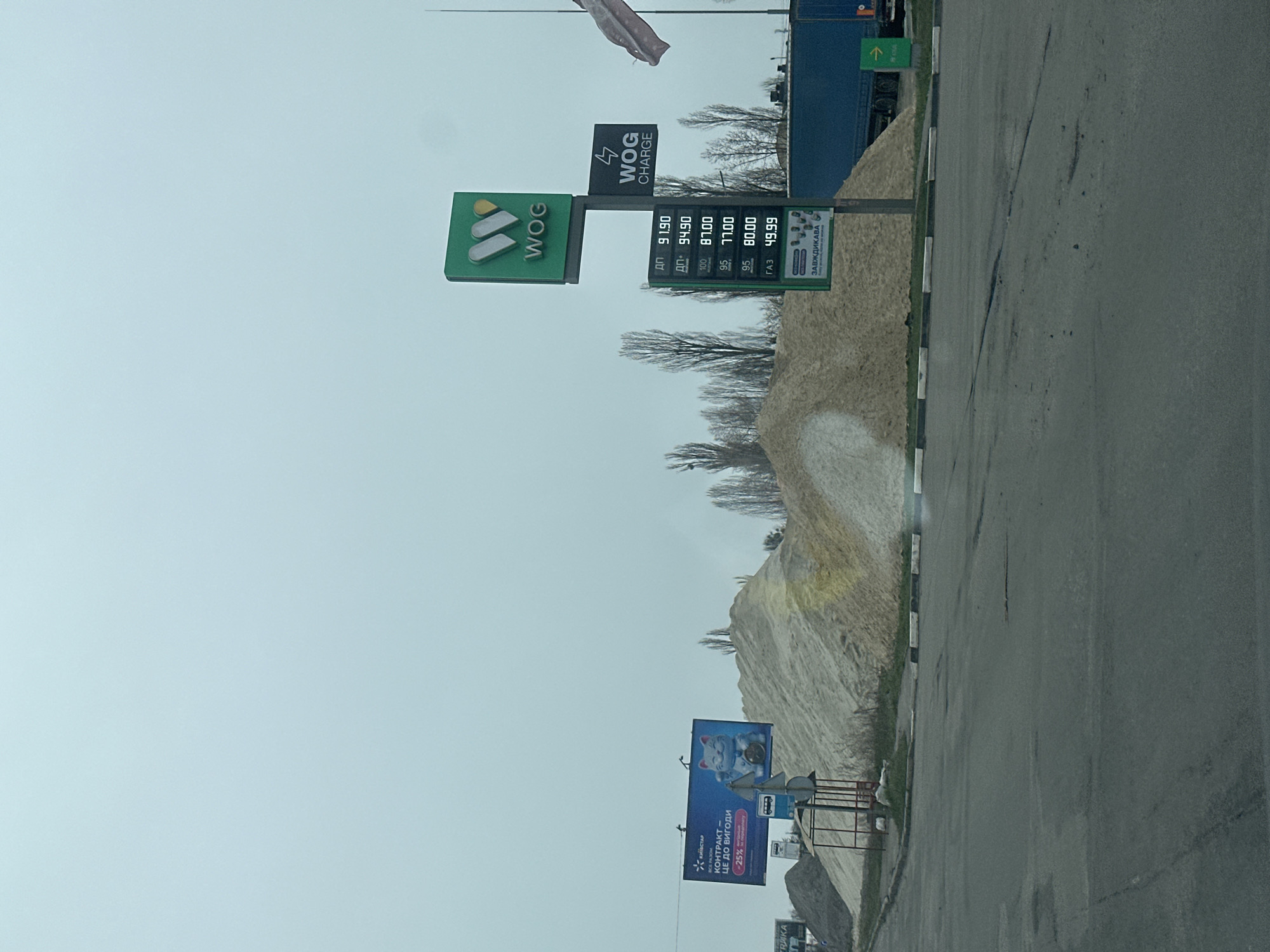 Here's a concise description of the image:
The image depicts a roadside scene, potentially in Kyiv, Ukraine, given the presence of signs written in Ukrainian. A WOG gas station is prominently featured, with listed fuel prices. A billboard with a Maneki Neko lucky cat can be seen in the background. The weather appears overcast. The scene shows an urban environment with roadside structures, billboards, and infrastructure.