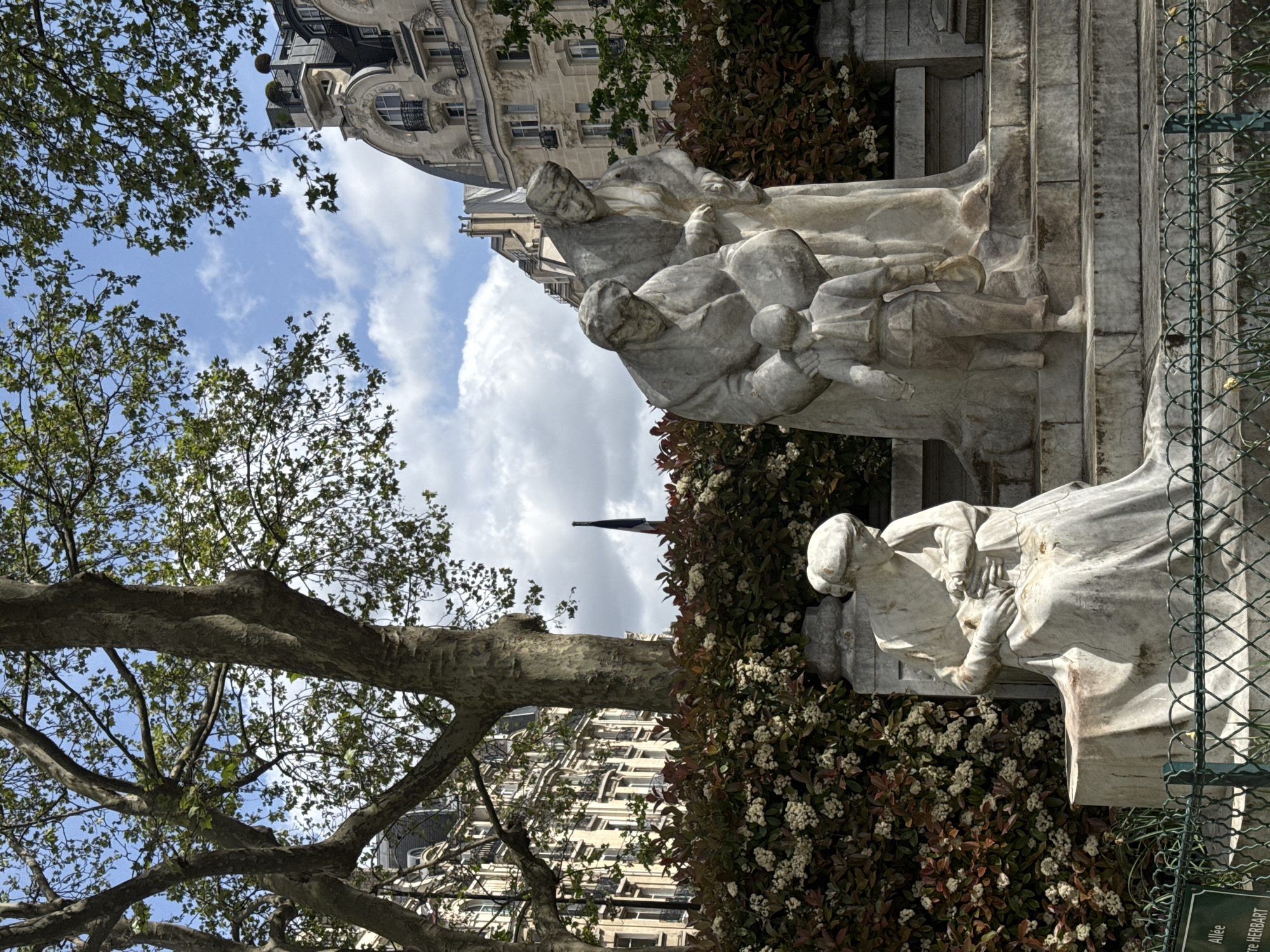 This image captures a serene scene in a Parisian park or garden, featuring a prominent group of stone statues under a bright sky. In the foreground on the left, the sturdy trunk and branches of a large tree with green foliage frame the view.
The central focus is a collection of light-colored marble or stone statues, depicting several human figures arranged on a tiered pedestal. The figures appear to be a family group, with adults and possibly children, rendered in a classical or realistic style. They are situated amidst manicured bushes with dark green and reddish leaves, dotted with small white flowers.
In the background, characteristic Parisian architecture is visible, likely Haussmannian buildings with ornate facades, balconies, and dormer windows, indicating an urban setting. Above the buildings and through the trees, a bright blue sky with scattered white clouds suggests a clear, sunny day, likely morning or afternoon due to the quality of light and shadows.
On the far right, a green metal fence runs vertically, and a small green sign with white text is partially visible at its base. The legible text on the sign reads "Allée Pierre HERBART," which translates to "Pierre Herbart Path," confirming the location as a public park or garden in Paris. The overall atmosphere is tranquil and picturesque, blending natural elements with urban architecture and art.