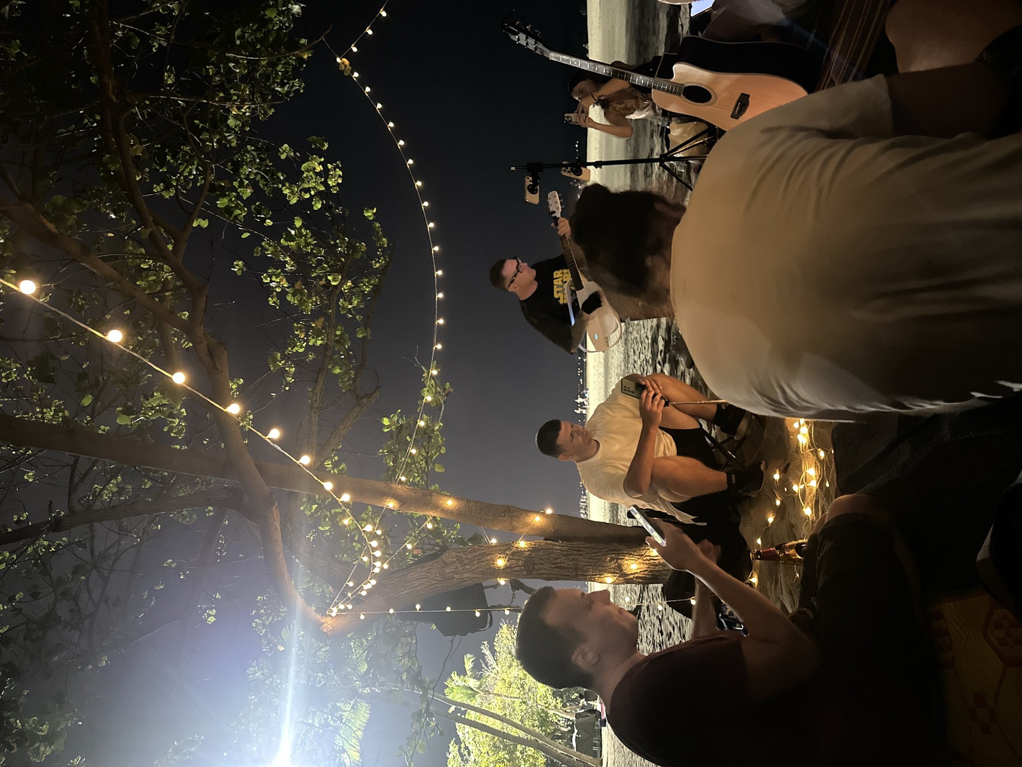 This nighttime photograph captures a casual outdoor gathering, likely a music session, set on what appears to be a beach or sandy area under a dark sky in Pattaya City, Thailand. The scene is illuminated by numerous warm-toned string lights draped through large trees, casting a gentle glow on the surroundings and the people.
Several individuals, mostly men, are present. In the foreground, partially obscured, a person in a maroon shirt is holding a smartphone, looking towards the center of the gathering. Another man in a white T-shirt and dark shorts sits on the ground, also engrossed in his phone. The focus of the musical activity is a man in a black "STAR WARS" T-shirt and glasses, actively playing a white electric guitar. Behind him, another person is seen with an acoustic guitar, seemingly also engaged in music or recording with a phone. A microphone stand with phone mounts is positioned near the musicians, suggesting an intent to record or livestream.
The atmosphere appears relaxed and informal, with individuals sitting comfortably on mats or blankets on the sandy ground, surrounded by the soft illumination of the fairy lights. A brown bottle is visible near some of the lights on the ground. The background is a dimly lit expanse of what seems to be a shoreline, with faint lights of a distant city visible, reinforcing the coastal location. The lush foliage of the trees adds to the natural outdoor setting. The overall mood is one of an intimate, casual evening event with music and social interaction.