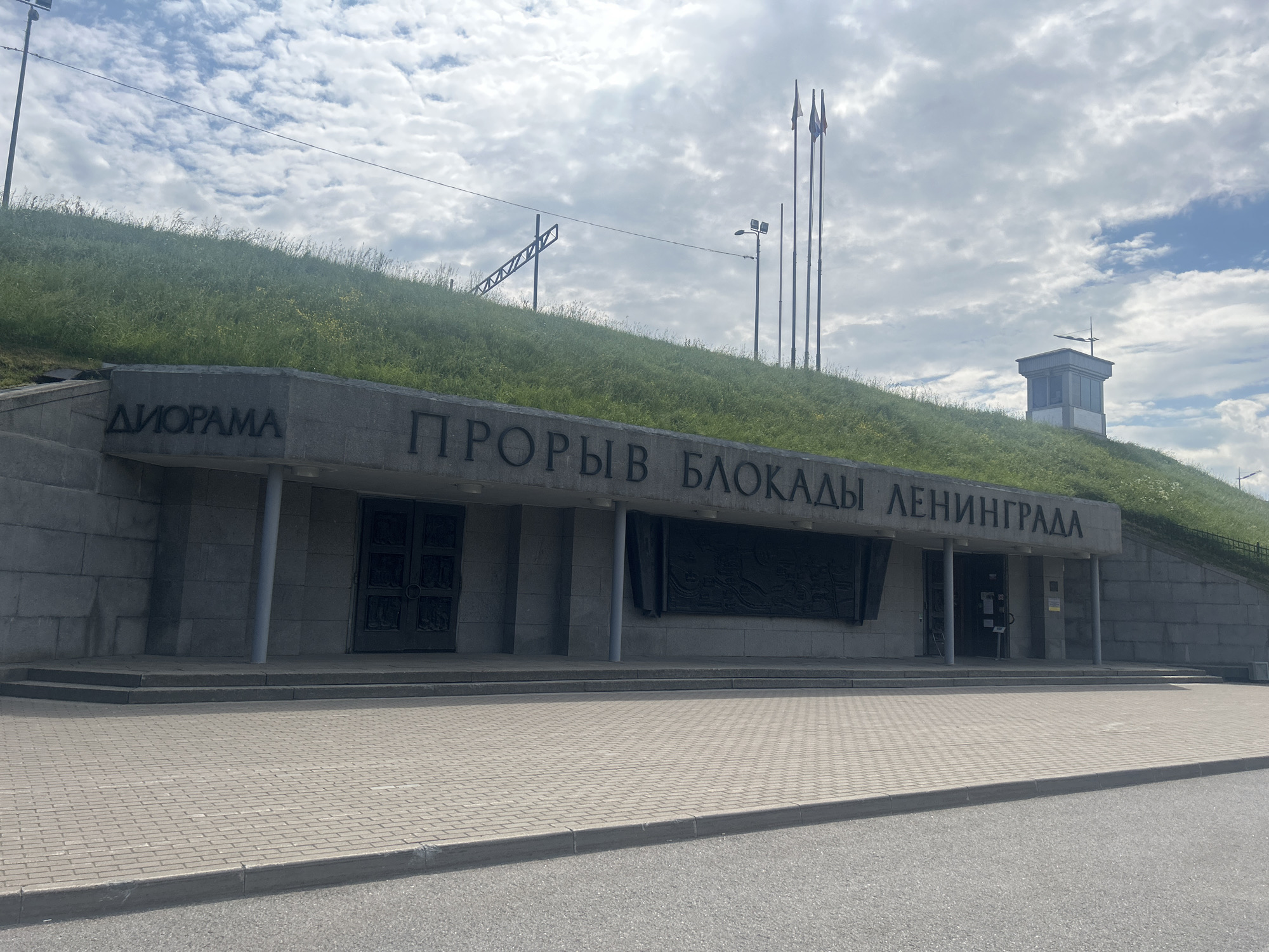 The image depicts the exterior of a building with a green, grassy embankment behind it. The building's facade is constructed of stone blocks. Above the entrance, large, dark, raised letters spell out "ДИОРАМА" and "ПРOРЫВ БЛОКАДЫ ЛЕНИНГРАДА" in Russian, translating to "Diorama" and "Breakthrough of the Siege of Leningrad." To the left of the entrance, there is a large, dark double door with ornate carvings. To the right of the entrance, there is a smaller single door. A large, dark, bas-relief artwork is mounted on the wall between the main entrance and the right-hand door. The building is supported by several concrete columns. The ground in front of the building is paved with light-colored brickwork. Behind the building, a grassy hill rises, dotted with various grasses and some yellow wildflowers. In the background, several flagpoles with flags are visible on the hill, along with a small, grey, modern structure with a pitched roof and a lantern on top. The sky is cloudy with patches of blue.