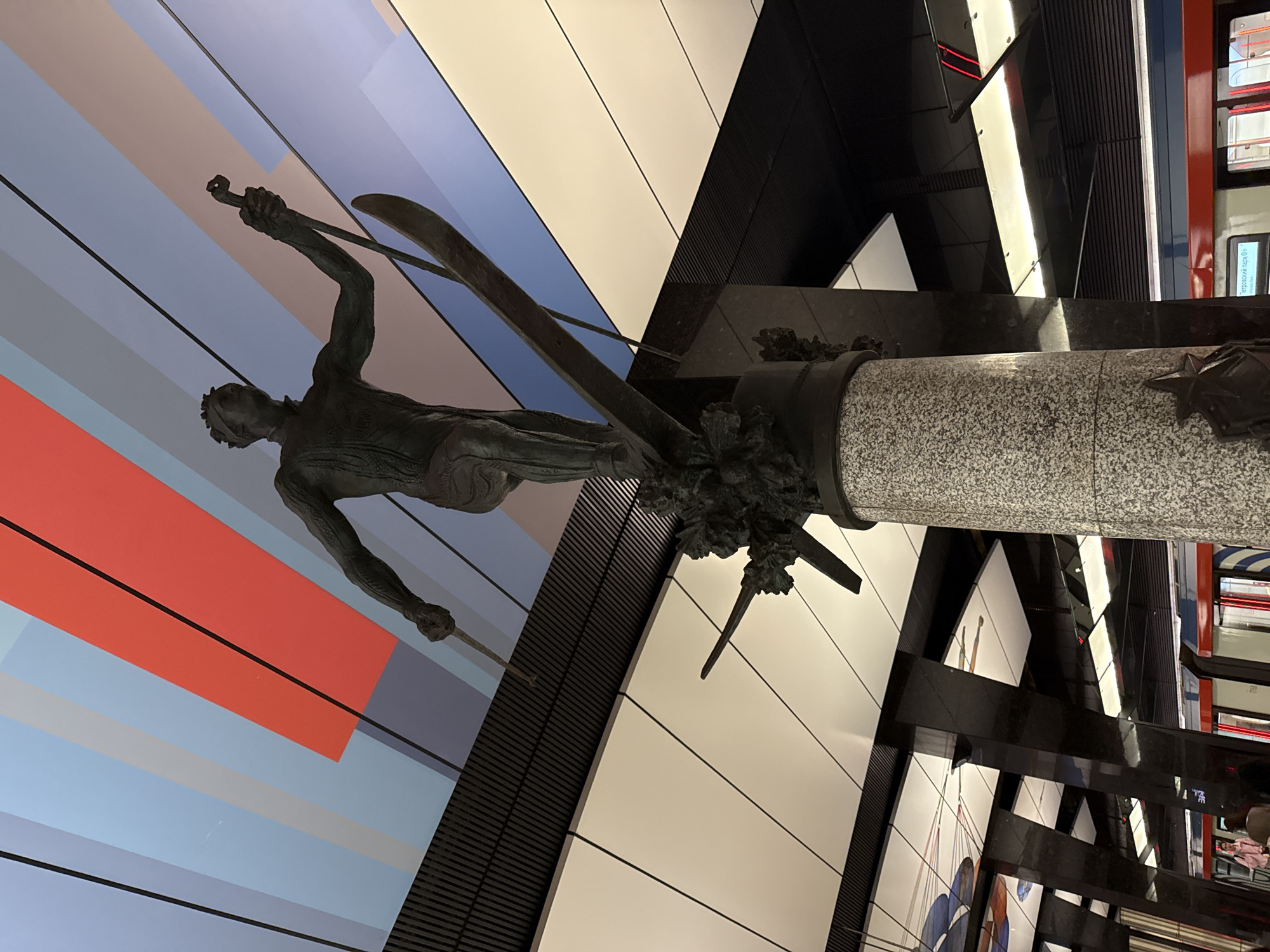 A statue of a skier stands in a Moscow Metro station. The station features a color block mural in shades of red, white, and blue. The statue sits atop a marble plinth adorned with bronze decorations. The background includes a train platform with a train visible.
