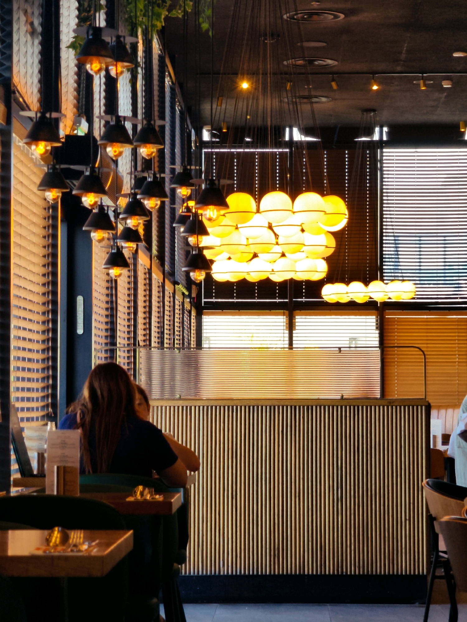 The image depicts the interior of a restaurant in Netivot, Israel, with a focus on the decorative lighting and seating areas. Overhead, a cluster of pendant lights with black shades and exposed yellow bulbs hang at varying heights, casting a warm glow. Further back, a larger, more elaborate light fixture composed of multiple spherical, frosted glass globes also illuminates the space with a soft, yellow light.
In the foreground, two individuals are seated at a table. The person in the foreground is seen from behind, with long, brown hair, wearing a dark blue top. They are seated at a wooden table with place settings, including cutlery. Another person is partially visible next to them.
The restaurant features a modern design with vertical wooden slats forming a partition or counter in the middle ground, separating the seating area from what appears to be a service or reception area. Behind this partition, horizontal blinds cover windows, allowing diffused light to enter the space. The ceiling is dark and industrial, with visible lighting fixtures and ductwork. The overall ambiance is warm and inviting, likely intended for evening dining.