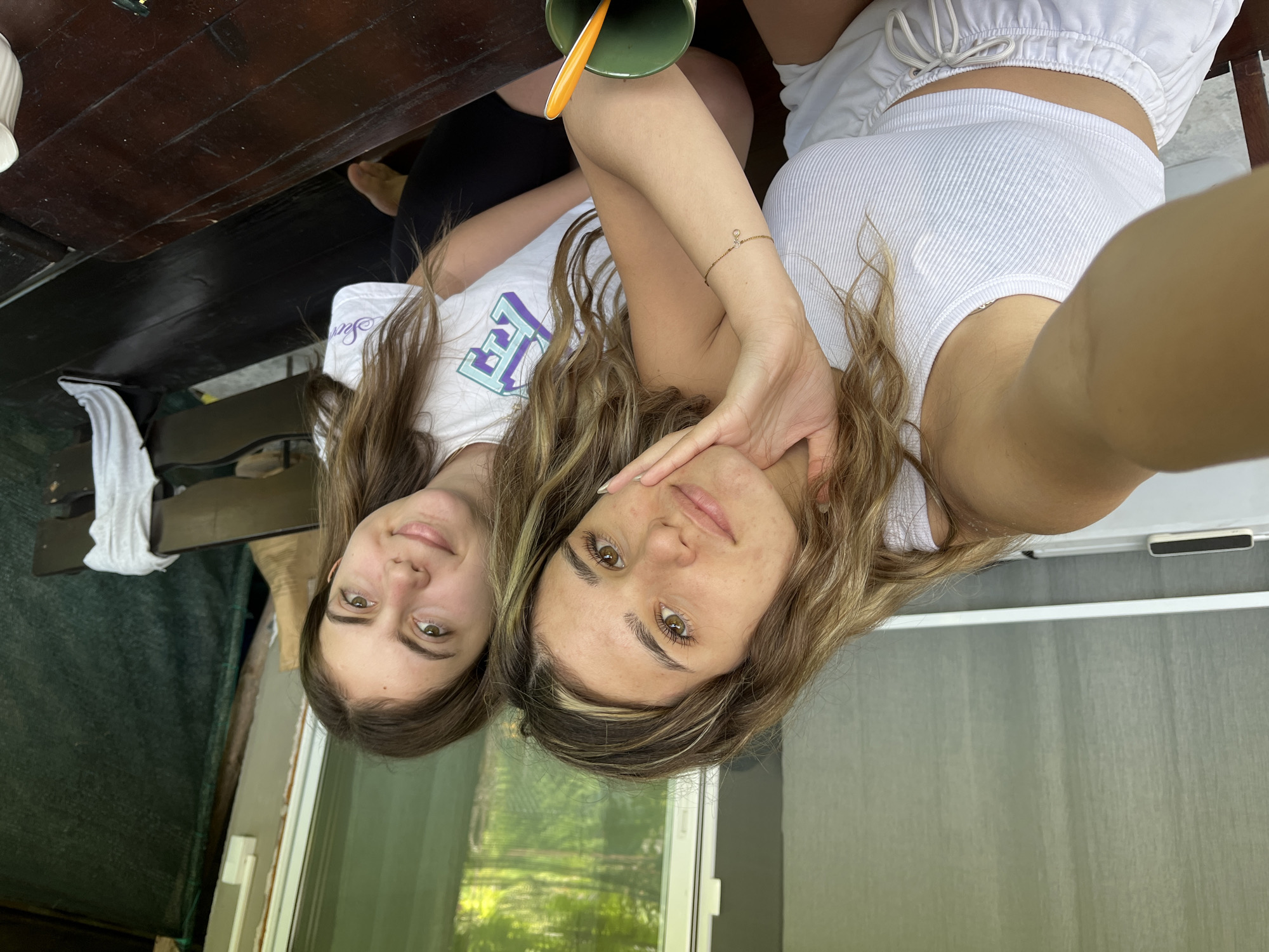 Two young women are posing for a selfie outdoors, possibly on a porch or veranda overlooking a natural landscape. The woman on the left is closer to the camera and is wearing a white ribbed tank top and white shorts. She has long, wavy brown hair and is resting her chin on her hand. The woman on the right is wearing a white t-shirt with a graphic design and black shorts. She has long brown hair and is smiling at the camera. In the background, there is a wooden table, a green mug with an orange spoon, and a dark green tarp. The location is identified as Tsarevo, Bulgaria.