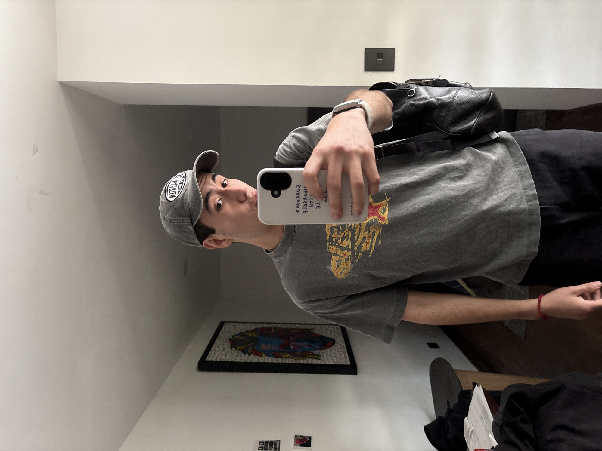 A young adult male, wearing a grey t-shirt with a graphic print, a grey baseball cap, and black pants, is taking a mirror selfie in what appears to be an indoor residential setting. He is holding a white smartphone in his right hand. A black backpack is slung over his left shoulder. The background includes a wall with a framed artwork featuring a colorful, stylized portrait, and a doorway leading to another room. The lighting suggests an interior space, likely a hallway or room within a house. The man is standing, facing the mirror, with a neutral expression. The scene does not contain any specific indicators of the city of San Bernardino Tlaxcalancingo, Mexico.