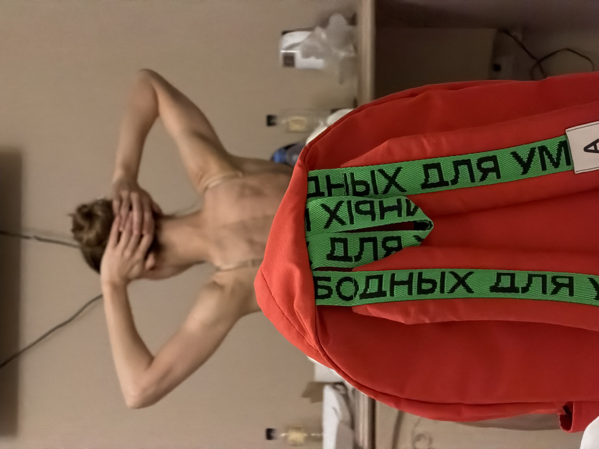 In Zelenogradsk, Russia, a person is seen from behind, facing away from the camera, with their hands resting on their head. They are wearing a strapless garment. In the foreground, a red backpack is visible, featuring a green strap with black lettering in Russian that reads "ДЛЯ ВСЕХ" (for everyone). The background suggests an indoor setting with furniture and a shelf.