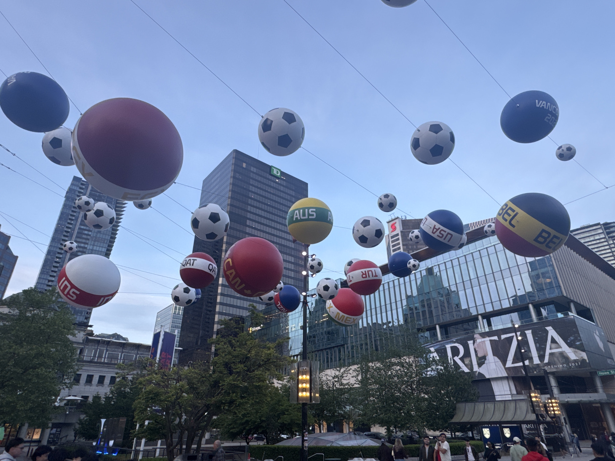 The image captures a street scene in Vancouver, Canada, decorated for a major sporting event, likely related to soccer. Several large, spherical decorations representing countries involved in the competition are suspended on wires above the street. The spheres are colored and labeled with country codes, like "AUS," "QAT," "CAN," "USA," "BEL," "TUR," and "MEX." Many standard soccer balls are also hanging on the wires between the larger country-themed spheres.
In the background are tall buildings, including one with a TD logo. A large "ARITZIA" sign is visible on another building, with the phrase "OPENING 2027." A street sign reads "Howe St."
Below the decorations, people are walking around, suggesting a busy public space. There are trees and street lights among the pedestrians, creating a lively and engaging urban environment. The sky is overcast, and the general lighting suggests either early morning or late afternoon.