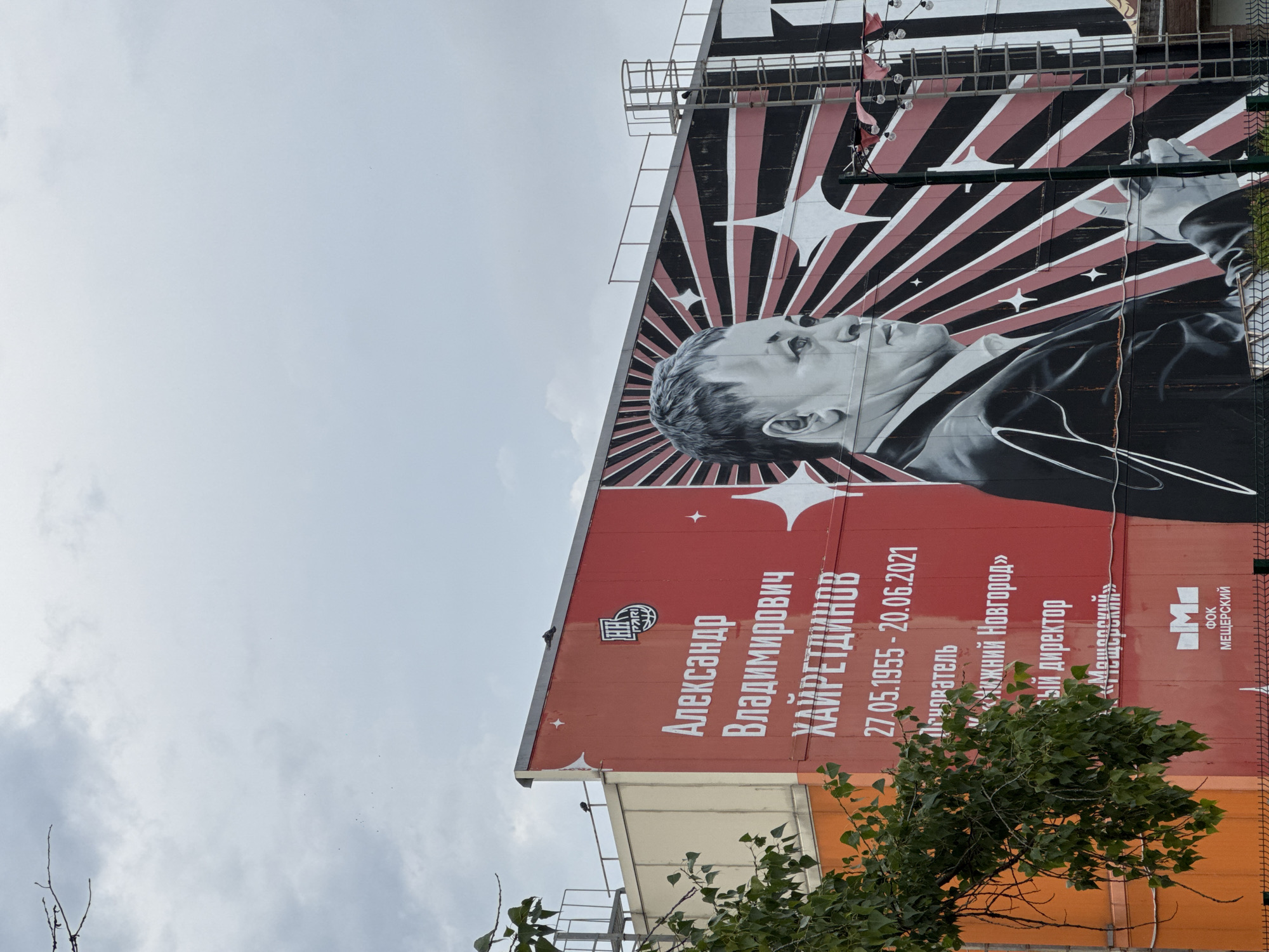 A large mural of Aleksandr Vladimirovich Khayretdinov is painted on the side of a building in Nizhny Novgorod, Russia. The mural features a portrait of Khayretdinov in a suit, with a stylized background of radiating black and red lines and white stars. Text below the portrait includes his name, birth and death dates (27.05.1955 - 20.06.2021), and his positions: "председатель" (chairman) of Nizhny Novgorod and "генеральный директор" (general director) of Meshchersky. The building itself is a large, modern structure with an orange section on the left and a red section on the right where the mural is located. There are metal railings and a ladder visible on the side of the building. A tree with green leaves partially obscures the left side of the mural. The sky above is overcast with grey clouds. The mural is located at the FOK Meshchersky.