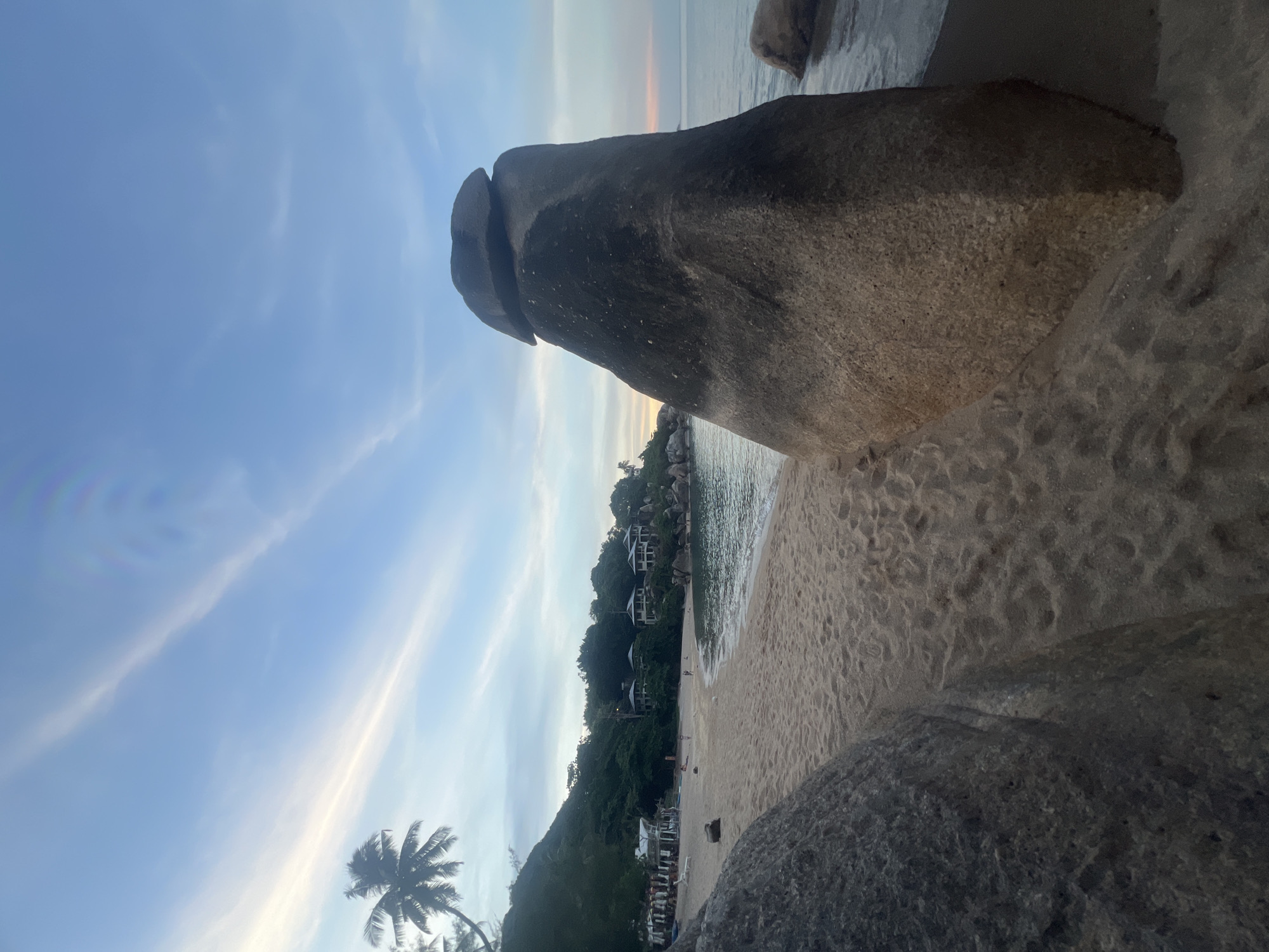 This is a vertical, eye-level, medium shot of a rocky beach on Ko Samui District, Thailand, during the late afternoon or early evening. The foreground is dominated by a large, dark-gray boulder on the left and a massive, light-brown boulder with a flat, stacked capstone on the right. The sand beach curves gently from the foreground towards the background, where a lush green hill dotted with multi-story buildings rises. A calm, turquoise sea laps at the shore, and a few small waves are visible. The sky is a soft blue with thin, wispy clouds, some tinged with orange from the setting sun. A single palm tree is visible on the left side of the image, its fronds silhouetted against the sky. A few small figures are scattered across the beach, and some lounge chairs and umbrellas are visible near the water's edge in the distance. The overall atmosphere is tranquil and serene.