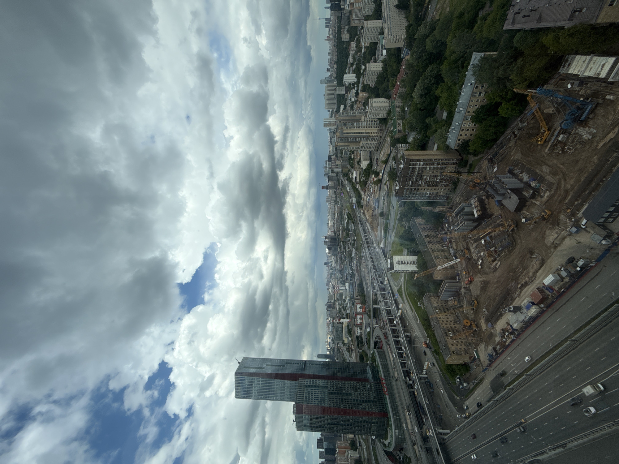 View from a high vantage point overlooking a city with a large highway. The clouds fill the left side of the frame and the city buildings fill the right. A modern skyscraper is visible in the lower left. Construction appears to be ongoing in the central right of the image. The highway stretches away into the distance. Based on the urban layout and general appearance, it is likely the city of Khimki in Russia.