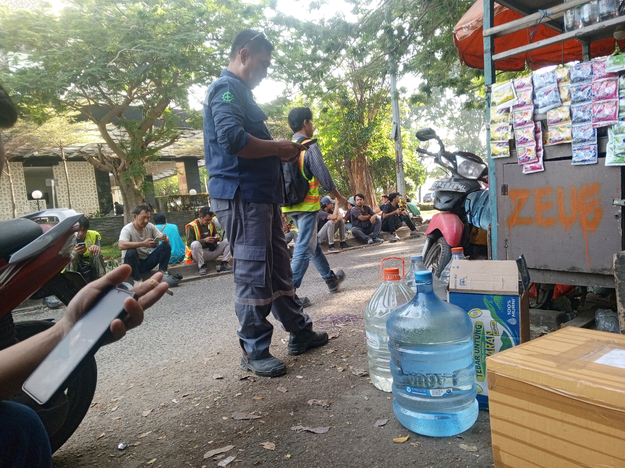 In the city of South Tangerang, Indonesia, an outdoor scene unfolds. A man in a dark blue uniform, with a green emblem on his left breast pocket and reflective strips on his pants, stands holding a mobile phone in his left hand and a lit cigarette in his right. He is positioned on a paved area with scattered leaves and debris. Behind him, another person in a yellow and black reflective vest walks away, carrying a backpack. Several other individuals are seated or standing in the background. To the right, large water cooler jugs are visible, one clear and one blue, placed on the ground. A red-orange awning is stretched over a makeshift stall laden with various packaged goods, likely snacks or beverages. A motorcycle is parked to the left, partially obscuring the view. Graffiti spelling "ZEUS" is spray-painted on a grey metal surface behind the stall. The environment suggests a commercial or service area, possibly a roadside stall.
