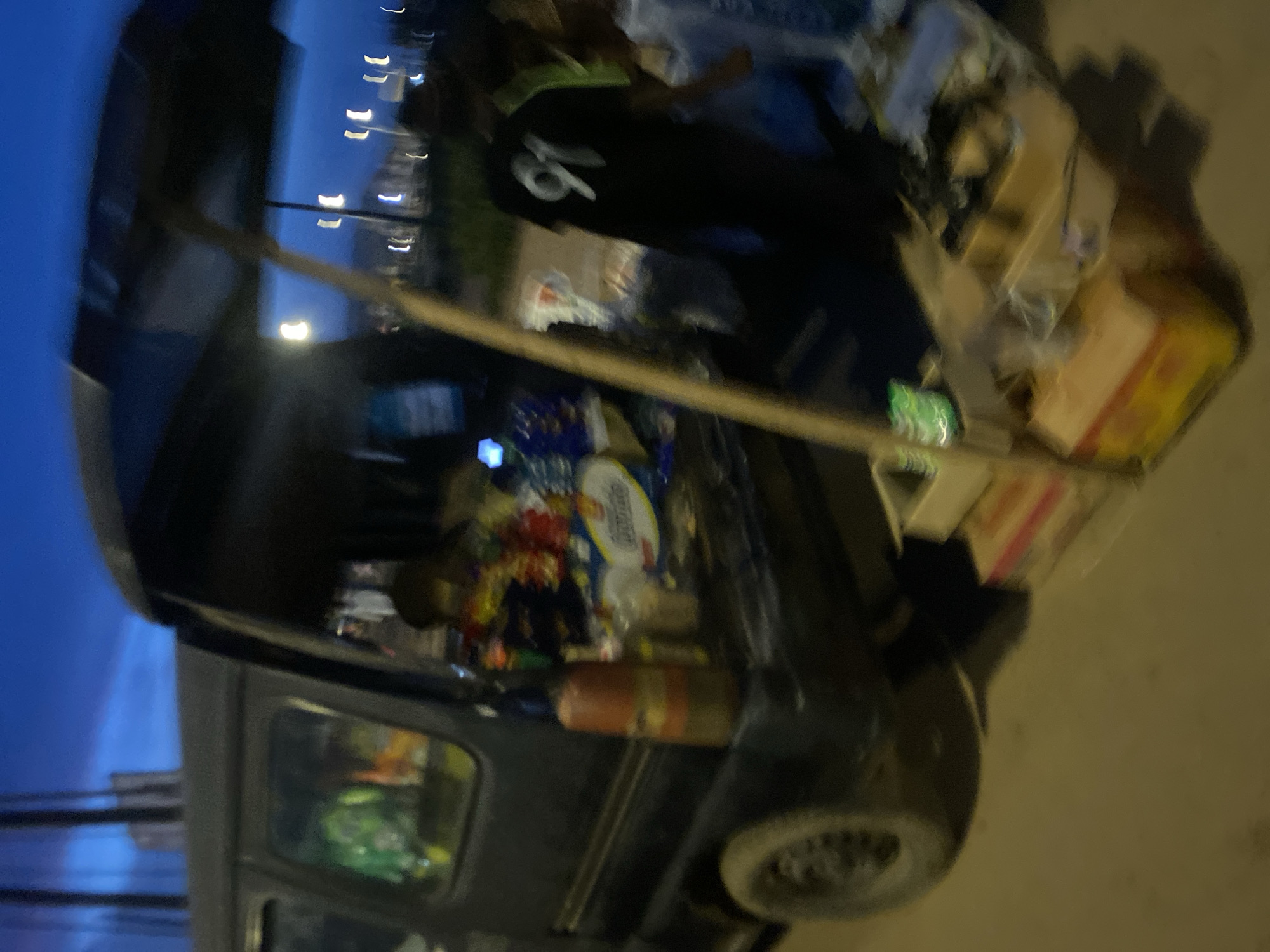 The image depicts a street vendor setup in Bukuru, Nigeria, likely at dusk given the dark blue sky. A dark-colored van is parked, with its rear door open, serving as a makeshift stall. The van is filled with various items, appearing to be snacks and drinks, with colorful packaging visible. A person, possibly the vendor, is partially visible inside the van. In front of the van, a display table is set up, also laden with goods. A person wearing a black t-shirt with the number "91" printed on the back stands near the table, facing away from the camera. Another person is partially visible to their right. A large umbrella is providing shade over the vendor's setup. The ground appears to be a paved road. Streetlights or distant building lights are visible in the background, suggesting an urban or semi-urban environment. The overall scene suggests a commercial activity taking place during the evening.