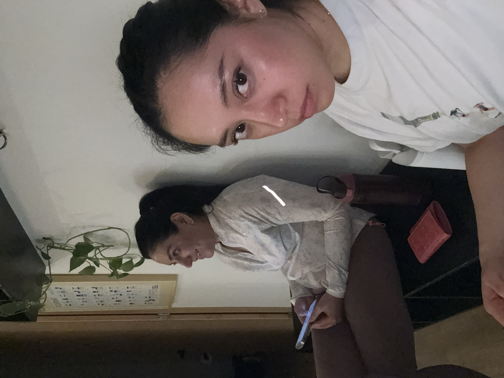 Two women are depicted in the image. The woman in the upper part of the image is wearing a white t-shirt. The woman in the lower part of the image is wearing a light-colored long-sleeved top. She is holding a smartphone. A potted plant and a framed picture are visible in the background. A red wallet and a bottle are also visible on a black surface. The setting appears to be indoors. The location is Puebla, Mexico.
