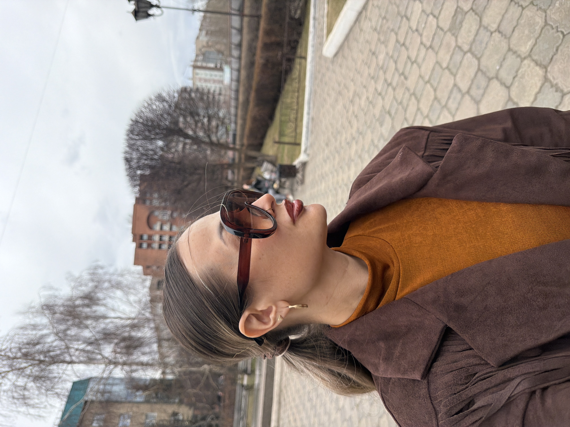 The image depicts a woman in profile, wearing sunglasses, a brown jacket, and an orange turtleneck sweater. She stands outdoors on a brick-paved area. In the background is a cityscape that includes a bare tree, buildings, and a lamppost. Based on the architectural style and urban layout, the location is likely in Krasnoyarsk, Russia.
