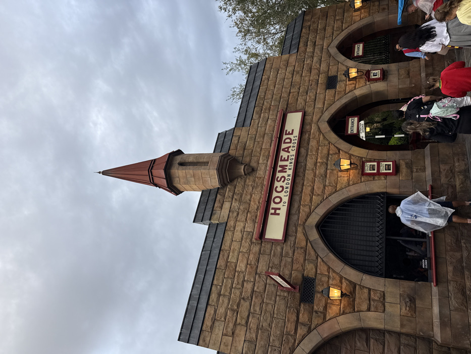 This image depicts the exterior of the Hogsmeade Station at Universal's Islands of Adventure, within The Wizarding World of Harry Potter in Orlando, Florida, United States of America. The architecture is a stone-block façade featuring multiple arched entryways and a prominent central turret with a reddish-brown conical roof, under an overcast sky.
A large sign centrally positioned above the arches reads "HOGSMEADE TO LONDON KINGS CROSS" in dark red lettering on a cream background. Below, three prominent arched openings are visible. The leftmost arch contains a barred window with a "WINDOW CLOSED" sign beneath it. A male individual wearing a translucent rain poncho over dark clothing stands facing this window.
The central archway is designated as an "ENTRANCE" and leads to an "ATTRACTION QUEUE," with multiple people entering. These include a female with long dark hair, wearing a dark top and carrying a light-colored backpack, and a young person in a bright red long-sleeved shirt. The rightmost archway also serves as an entrance or exit, where a female in a white shirt and pleated skirt walks towards the opening. A park employee in a red vest is partially visible behind her, and another individual in a rain poncho is visible to the far right. Multiple lanterns with illuminated orange light are mounted on the stone walls, flanking the arches. Green foliage is partially visible to the right of the structure.