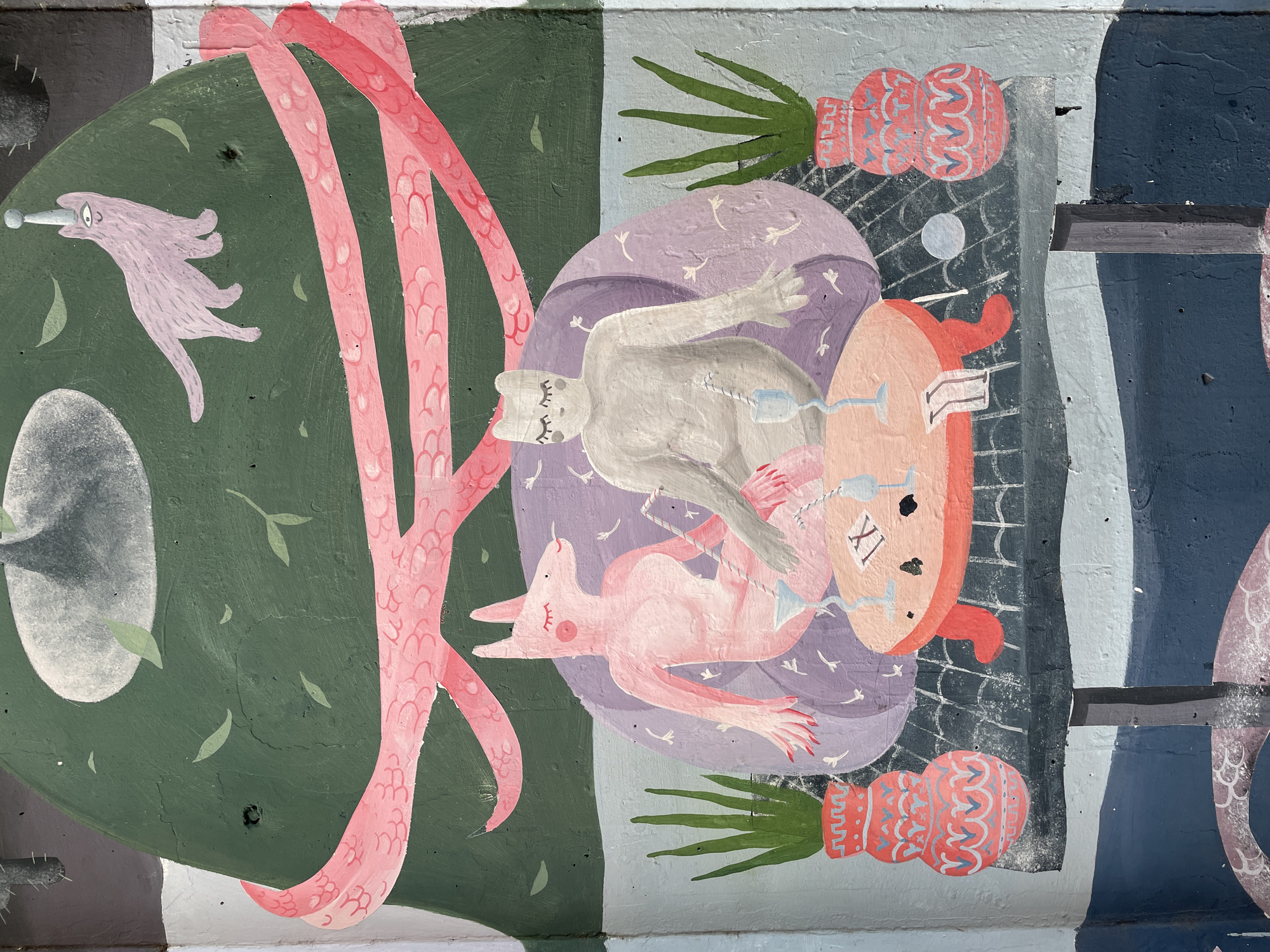 The image shows a section of a mural, likely an outdoor public artwork. The mural appears to be painted on a textured surface, possibly concrete. The scene is divided into distinct areas with different colors and elements.
On the left side, there's a green field with a stylized pink figure resembling a fantastical creature. A pink tentacle-like form rises vertically.
The central section contains a scene with reclining figures of light grey and pink animals in a purple field, possibly dogs, with glass objects floating above them.
To the right, there is a stylized clock face with Roman numerals and red ears, two ornamental vases, and green pineapple fronds. The background behind these elements is a mix of grey and black, and dark blue sections.
The mural is located in Podgorica, Montenegro.
